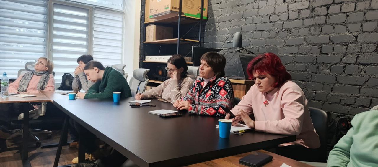 Група жінок сидить за довгим столом під час робочої зустрічі або навчання. Вони занотовують інформацію, на столі стоять паперові стаканчики. Тло — темна цегляна стіна та офісні меблі.