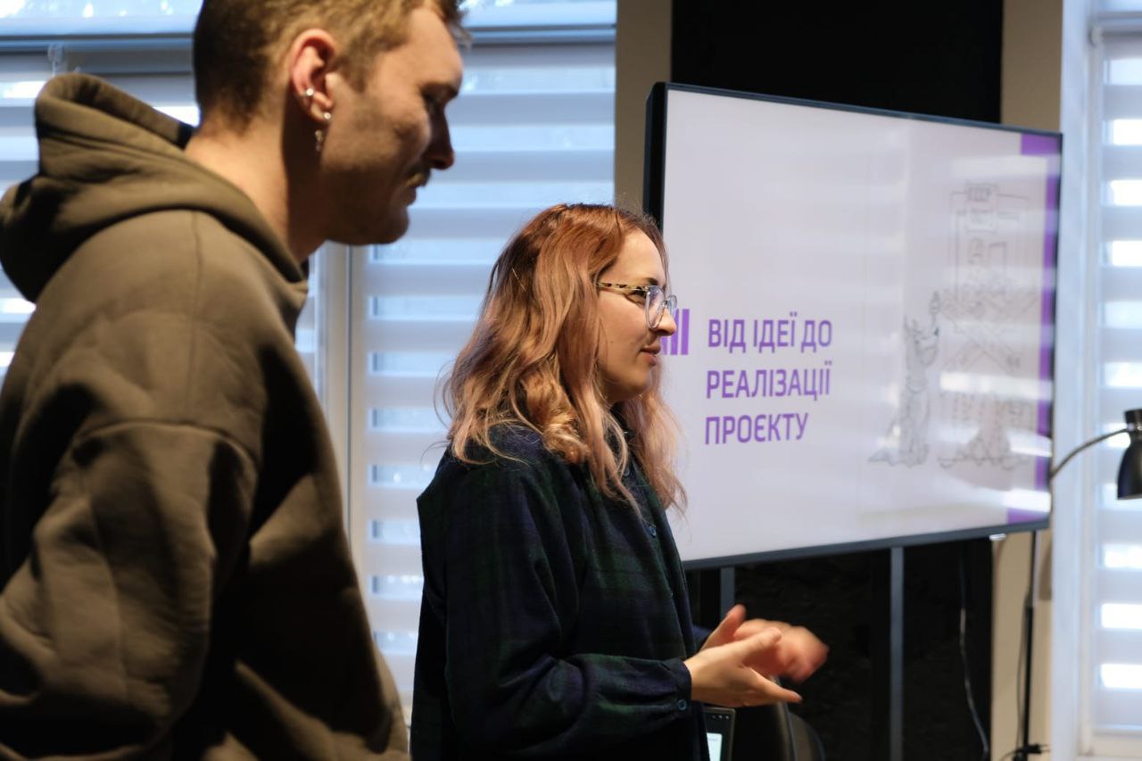 Двоє людей стоять біля екрана, на якому відображено слайд презентації з написом «Від ідеї до реалізації проєкту». Дівчина жестикулює, пояснюючи матеріал на фоні вікна з жалюзі.