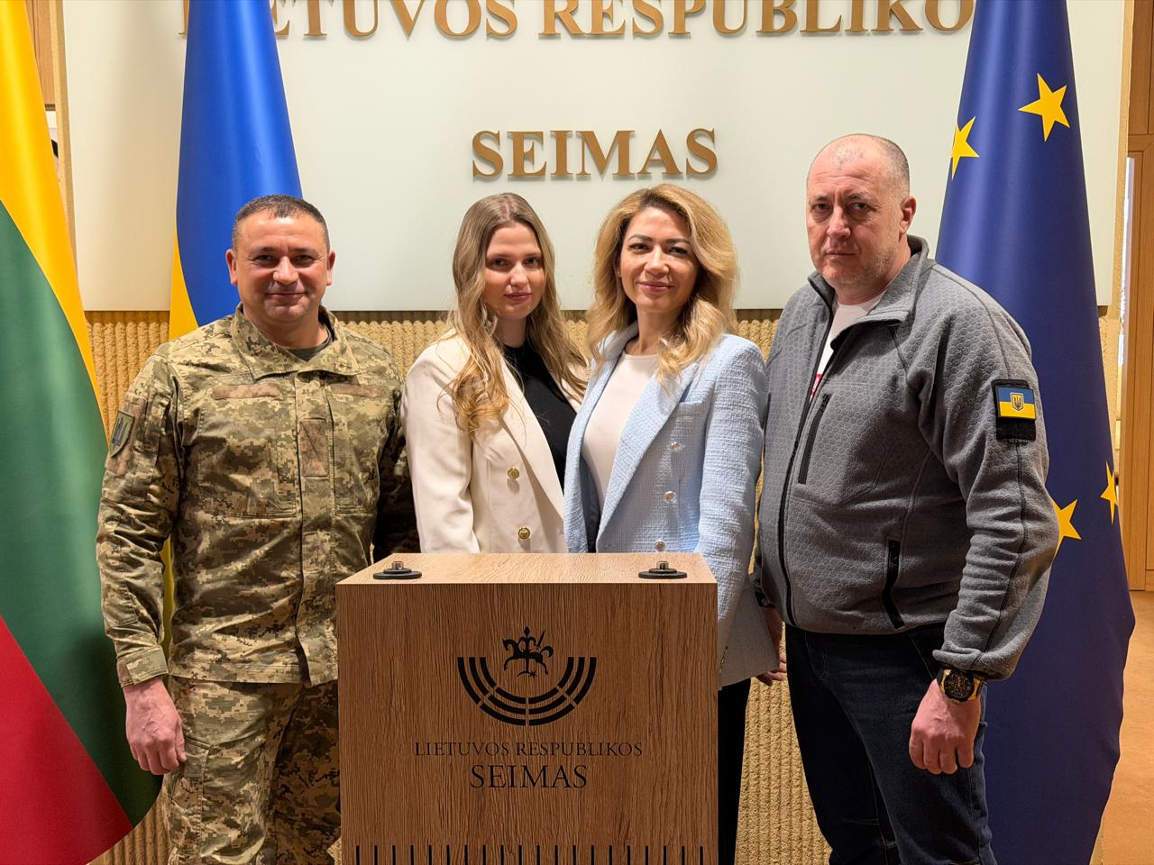 Чотири особи (двоє чоловіків та дві жінки) стоять за трибуною з гербом Сейму Литовської Республіки. Чоловік ліворуч одягнений у військову форму, чоловік праворуч — у спортивну кофту з прапором України на рукаві. Жінки в центрі одягнені у світлі піджаки. На задньому плані розташовані прапори Литви, України та Європейського Союзу.