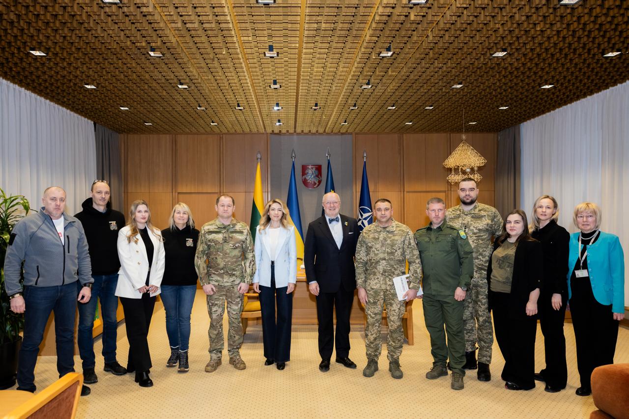 Офіційна зустріч групи осіб у цивільному одязі та військовій формі в офіційному залі. У центрі стоять жінка у світлому піджаку та чоловік у костюмі. На фоні — прапори Литви, України, НАТО та герб Литви під рельєфною дерев'яною стелею.