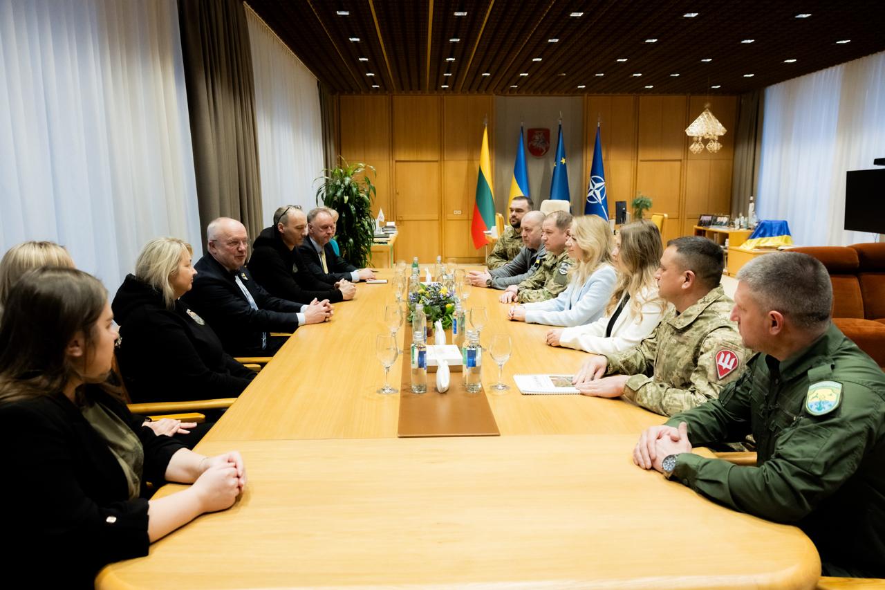 Дві групи людей у цивільному та військовому одязі проводять зустріч за довгим дерев'яним столом. Учасники сидять один навпроти одного в офіційному кабінеті. На задньому плані видно прапори Литви, України, Європейського Союзу та НАТО, а також герб Литви на стіні.