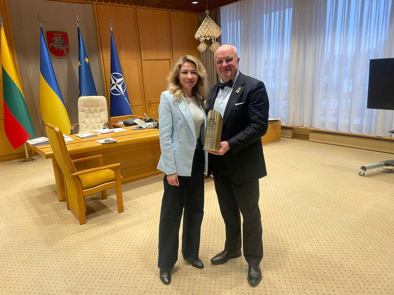 Жінка у світло-блакитному піджаку та чоловік у класичному костюмі з метеликом стоять у кабінеті. Чоловік тримає пам'ятну нагороду золотистого кольору. На фоні видно робочий стіл, прапори Литви, України, Європейського Союзу та НАТО, а також державний герб на стіні.