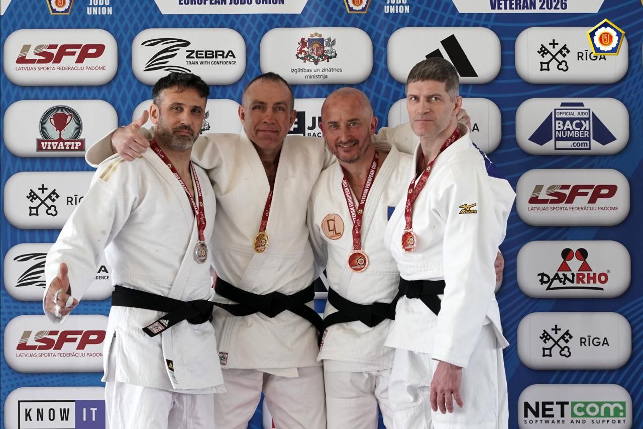 Чотири спортсмени-дзюдоїсти у білих кімоно з медалями на шиях стоять поруч, обійнявшись, на фоні брендованого банера з логотипами спортивних організацій та спонсорів.