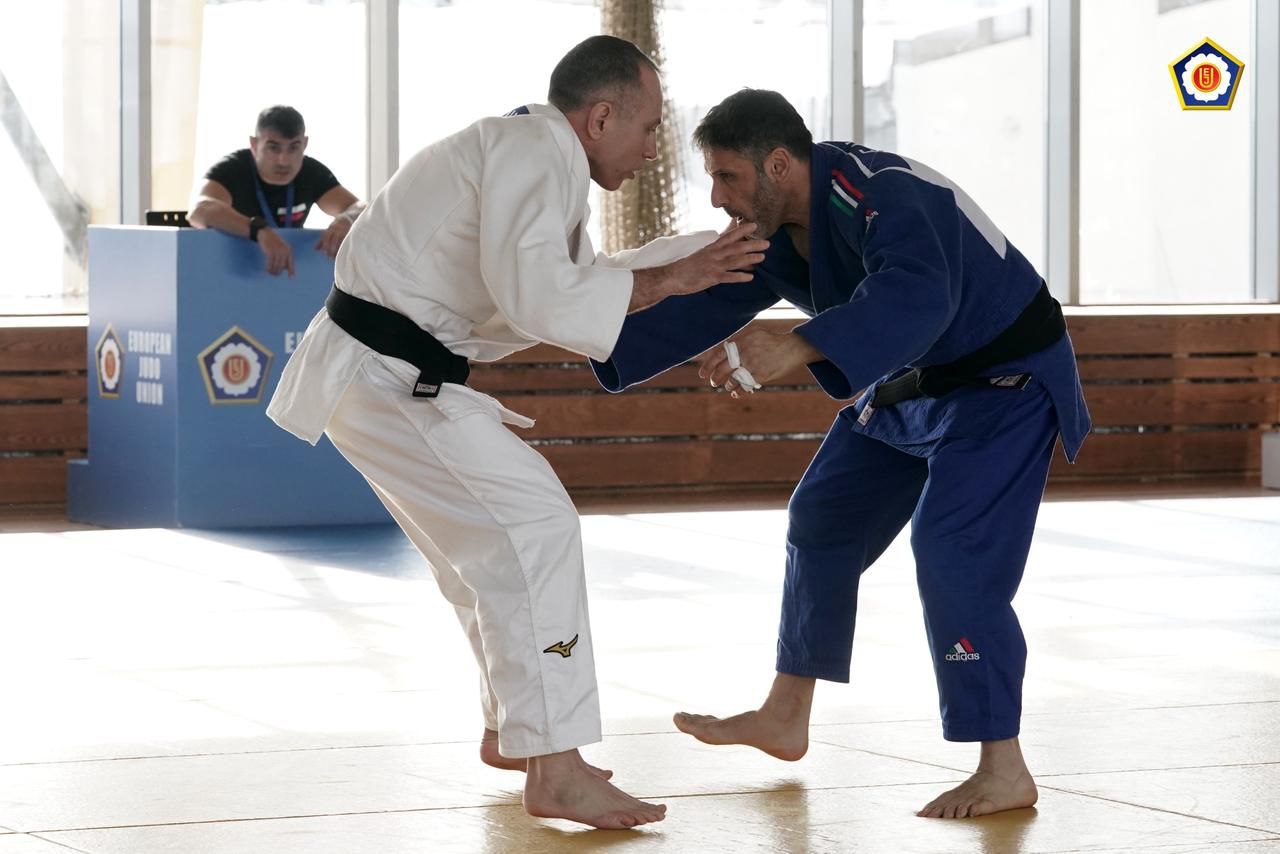 Двоє дзюдоїстів у білому та синьому кімоно змагаються на татамі в сонячному залі. На задньому плані за стійкою сидить суддя або організатор, а поруч на стіні розміщено логотип спортивної федерації.