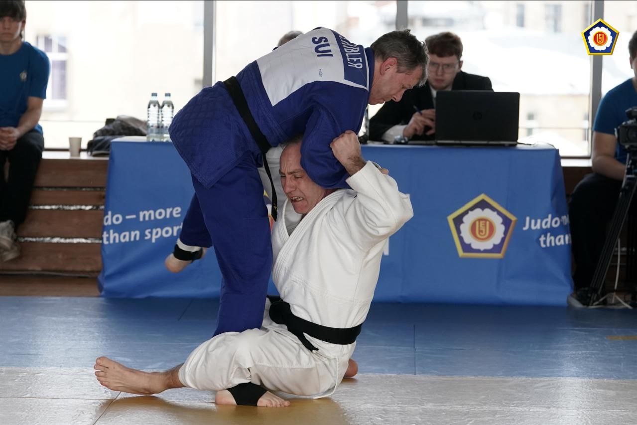 Двоє дзюдоїстів ведуть боротьбу на татамі під час змагань. Спортсмен у синьому кімоно виконує кидок через плече проти суперника у білому кімоно. На задньому плані за столом із ноутбуком сидить суддя, а поруч розміщено банер із логотипом європейського дзюдо та написом «Judo more than sport».
