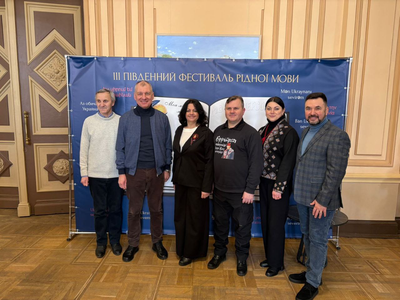 Група з шести осіб стоїть на фоні синього банера з написом «III Південний фестиваль рідної мови». Учасники одягнені у повсякденний та офіційно-діловий одяг, зокрема в українську вишиванку та світшот із цитатою Тараса Шевченка. Подія відбувається у приміщенні з класичним інтер'єром та високими дерев'яними дверима.