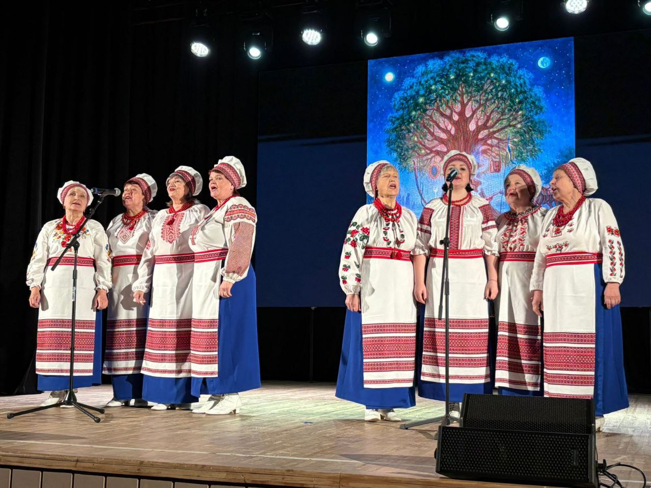 Ансамбль жінок у традиційному українському вбранні — вишитих сорочках, червоних намистах та очіпках — виступає на сцені. Вони стоять біля мікрофонів на фоні великого екрана із зображенням квітучого дерева під нічним небом.