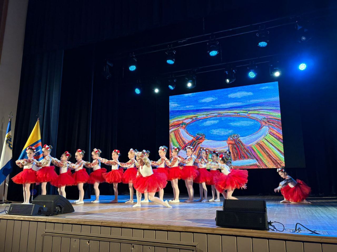 Дитячий танцювальний колектив у вишиванках та червоних спідницях-пачках виступає на сцені. Дівчата стоять у лінію, поклавши руки на плечі одна одній, на фоні великого екрана з яскравим пейзажем. Ліворуч на сцені встановлено прапор.