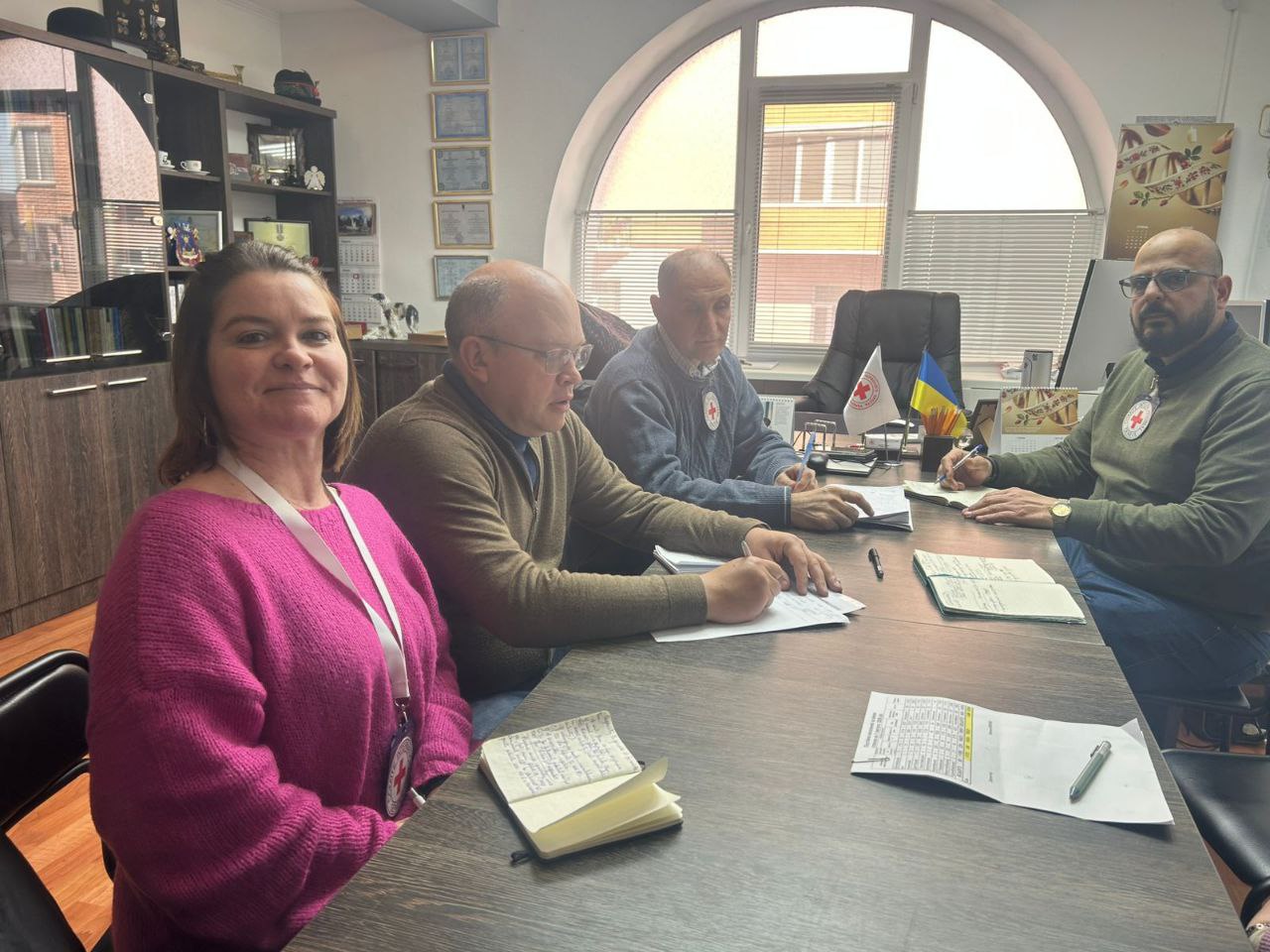 Чотири людини сидять за робочим столом в офісному приміщенні під час наради. На одязі присутніх видно емблеми Товариства Червоного Хреста. Учасники роблять записи в блокнотах; на столі лежать документи, а на задньому плані видно державний прапор України та грамоти на стіні.