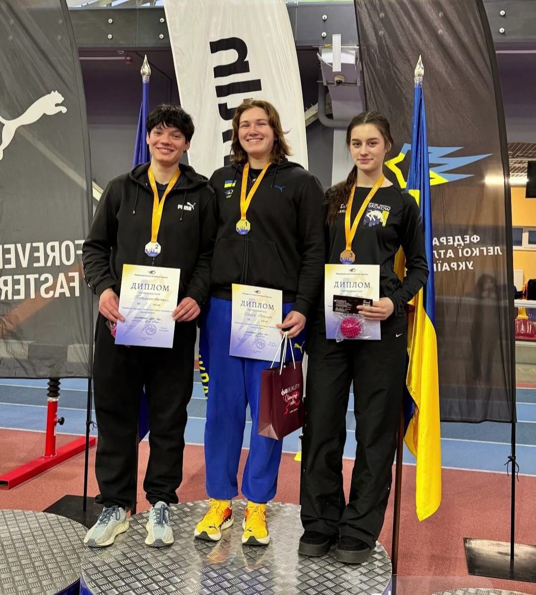 Три спортсменки стоять на п’єдесталі пошани в легкоатлетичному манежі. Кожна з них тримає диплом та має медаль на шиї (золоту, срібну та бронзову відповідно до місць). На фоні видно прапор України та спортивну символіку.