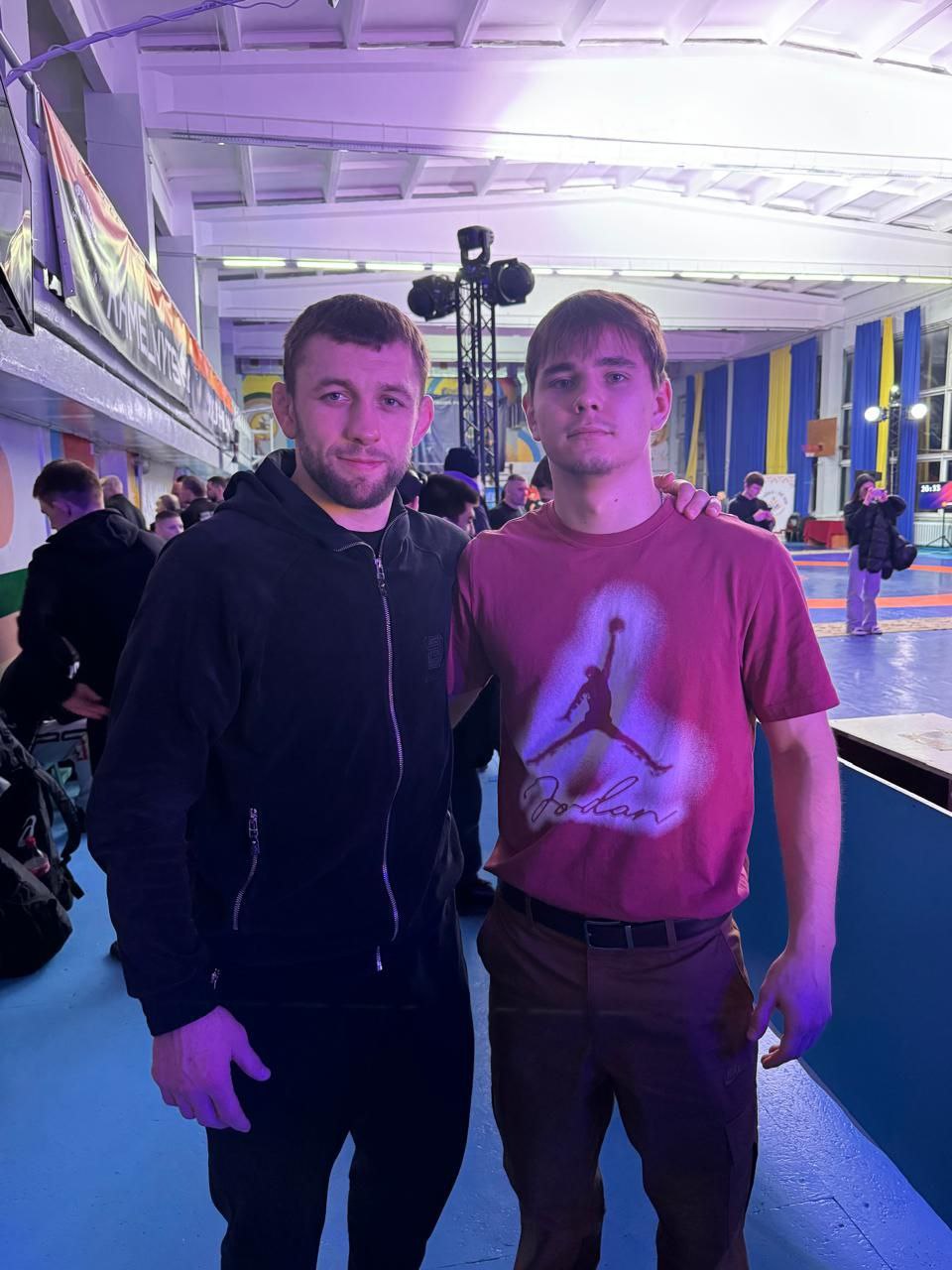Двоє спортсменів стоять поруч у спортивному залі після змагань. Один чоловік у чорній спортивній кофті, інший — у бордовій футболці. На задньому плані видно борцівський килим, прапори України та інших присутніх у залі людей.