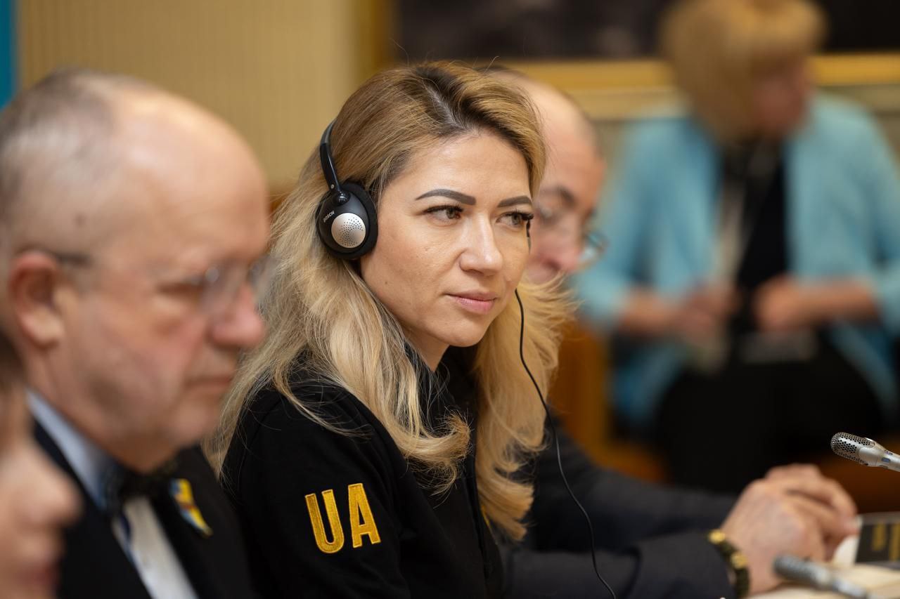 Портрет жінки з білявим волоссям у навушниках під час конференції. Вона сидить за столом поруч із колегами, на її одязі вишито напис «UA». Фокус на обличчі, задній план розмитий.