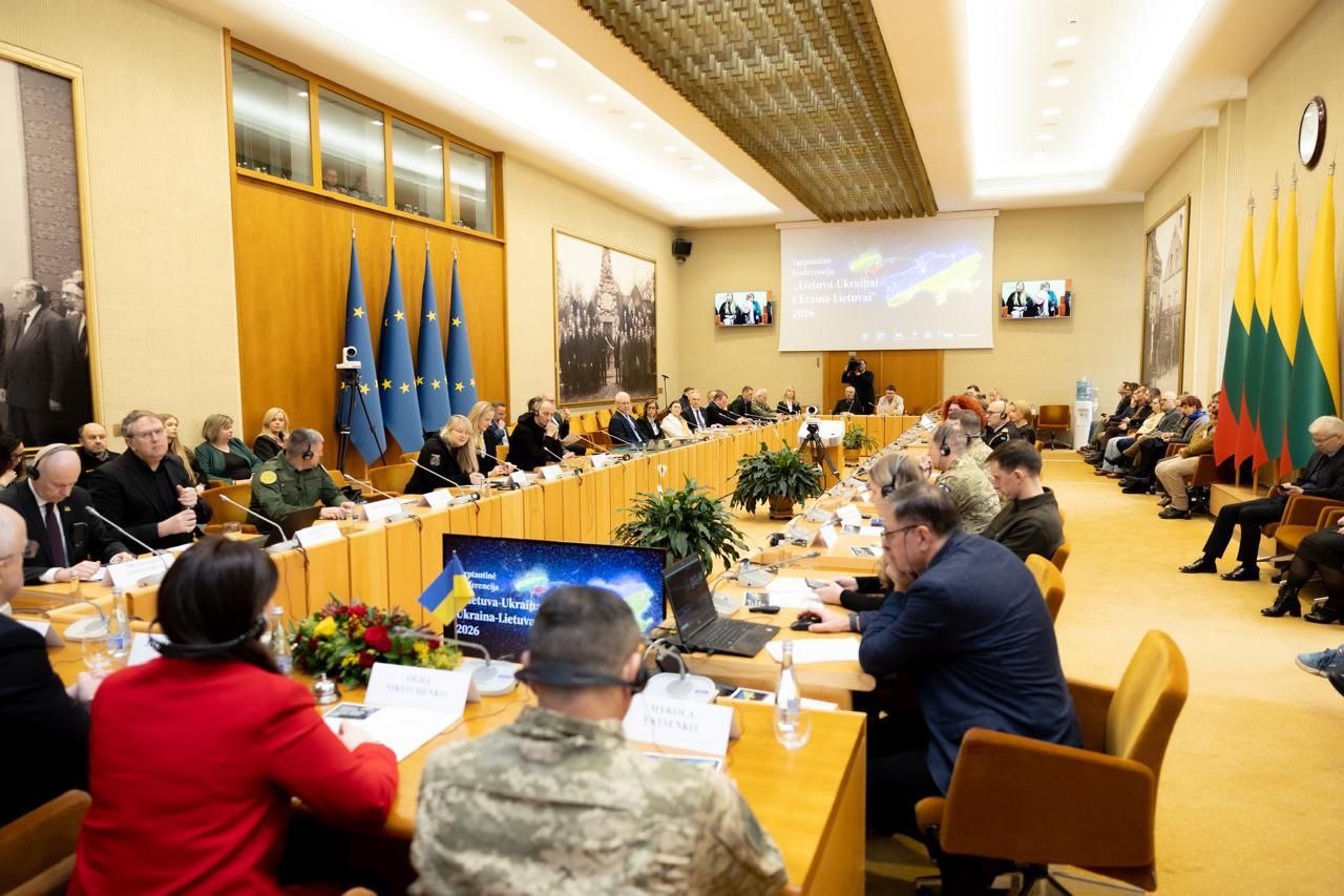 Загальний вигляд конференц-залу під час офіційної зустрічі. Учасники сидять за довгими столами, розставленими у формі літери «П». На передньому плані видно монітор, а на стінах — прапори Литви, Євросоюзу та великий екран із написом «Lietuva–Ukraina 2026».