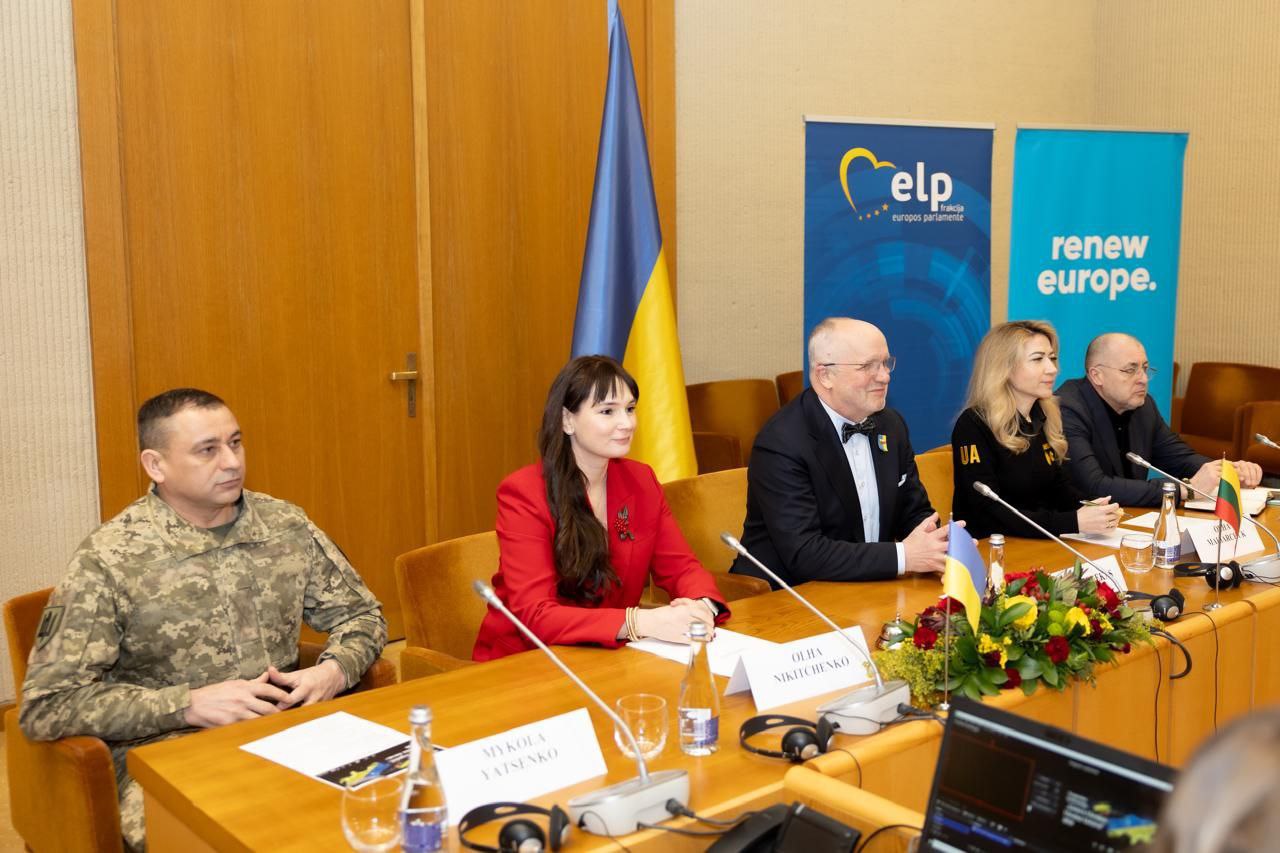 П'ятеро учасників офіційної зустрічі (військовий та четверо цивільних) сидять за круглим столом із мікрофонами та табличками з іменами. На фоні розташовані державний прапор України та банери з символікою «elp» та «renew europe».