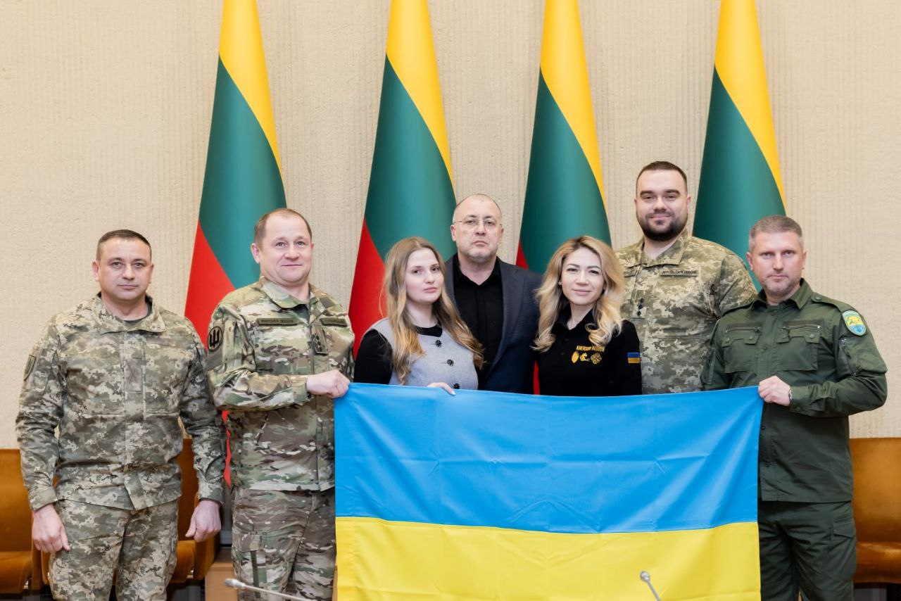 Офіційне фото групи з семи осіб (чотирьох у військовій формі Збройних Сил України та трьох у цивільному одязі), які спільно тримають розгорнутий державний прапор України. Вони стоять у ряд у світлому залі, а безпосередньо за ними розташовані три великі прапори Литви на флагштоках.
