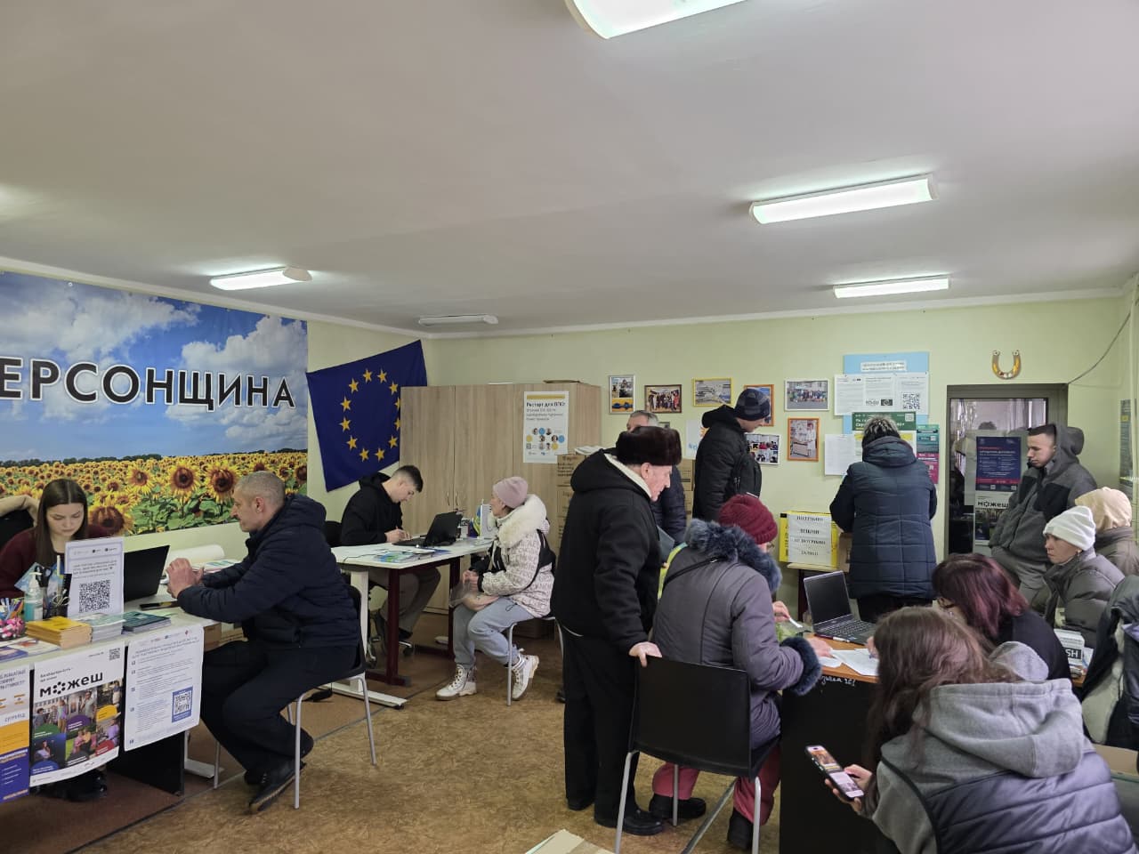 Люди в зимовому одязі перебувають у приміщенні гуманітарного або інформаційного центру, де на стіні розміщено плакат із соняшниками та написом «Херсонщина» і прапор ЄС.