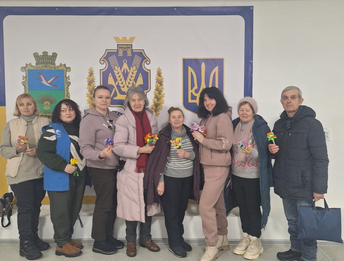 Група людей у зимовому одязі стоїть на фоні банера з гербами, тримаючи в руках яскраві саморобні квіти.