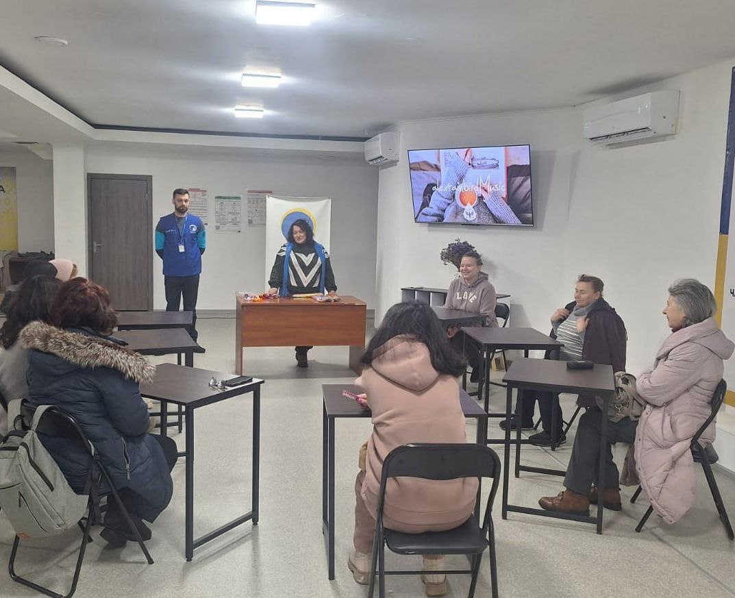 Учасники сидять за столами в аудиторії, слухаючи ведучу та чоловіка у синій уніформі.