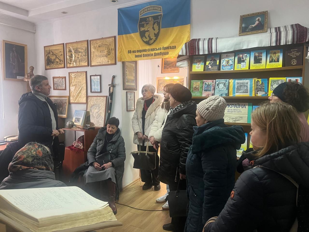 Група людей слухає екскурсовода в кімнаті, прикрашеній історичними картами, портретами та книжковою полицею. На стіні розміщено прапор 68-ї окремої єгерської бригади імені Олекси Довбуша.