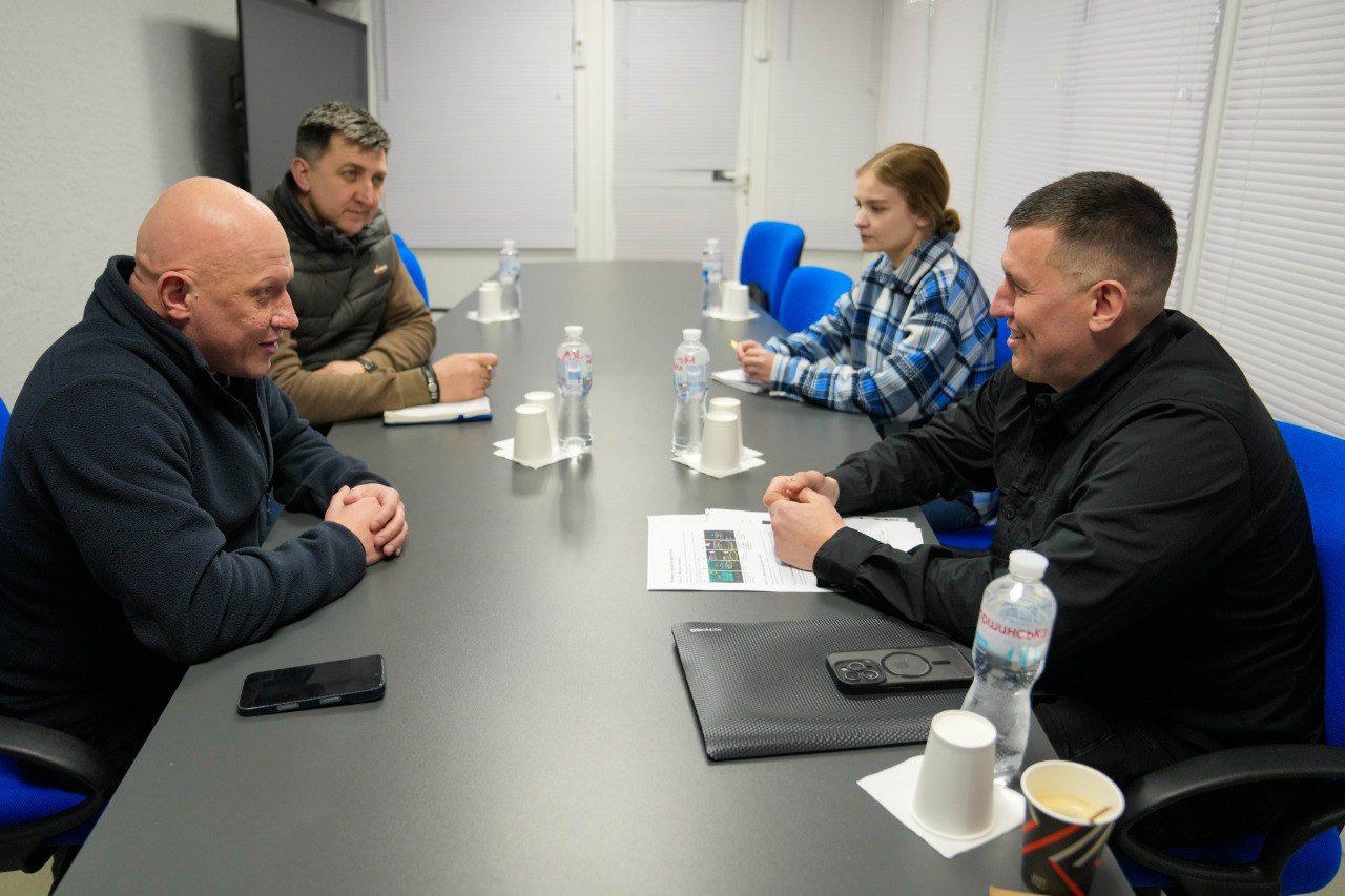 Четверо осіб проводять обговорення за довгим столом у світлому офісному приміщенні. На столі лежать документи, смартфони та стоять пляшки з водою. Учасники зустрічі зосереджено спілкуються в діловій атмосфері.