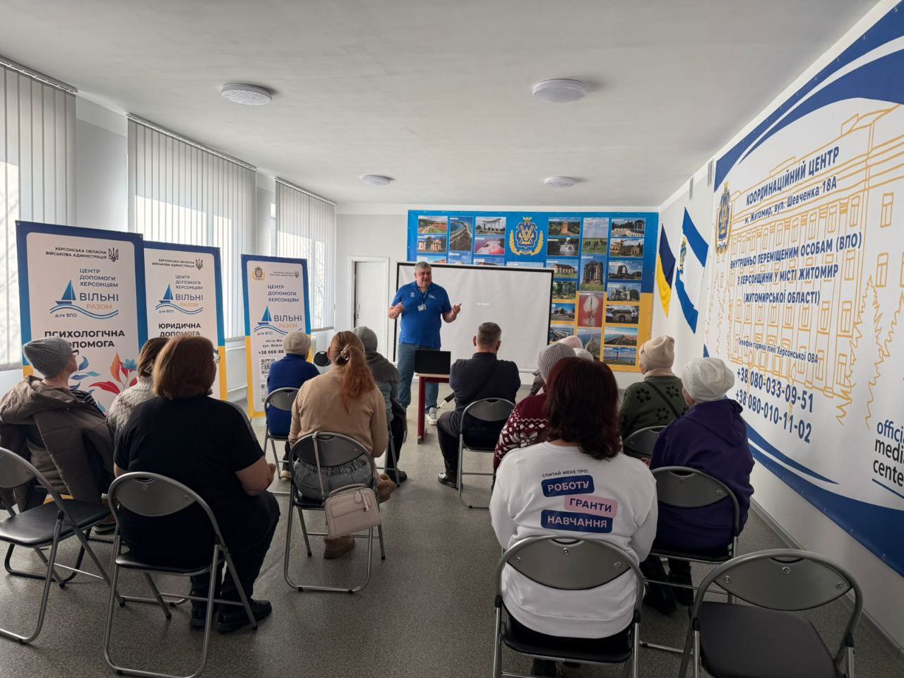 Презентація для переселенців у світлому залі з інформаційними стендами про психологічну та юридичну допомогу. На стіні розміщено банер Координаційного центру для херсонців у Житомирі.