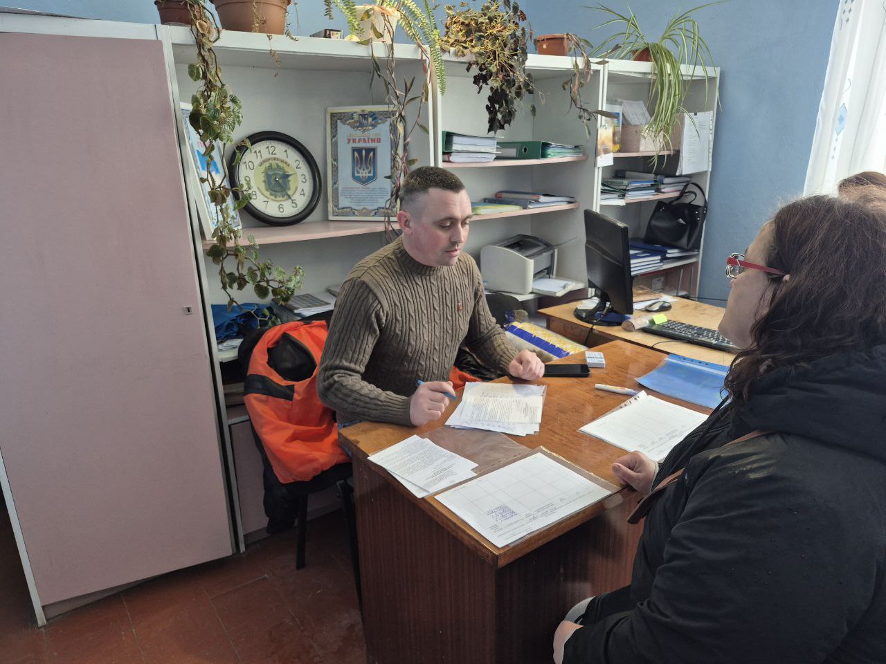 Чоловік у светрі проводить прийом відвідувачки в кабінеті. На робочому столі розкладено документи та бланки для заповнення. На фоні видно стелажі з папками, годинник та державну символіку України.