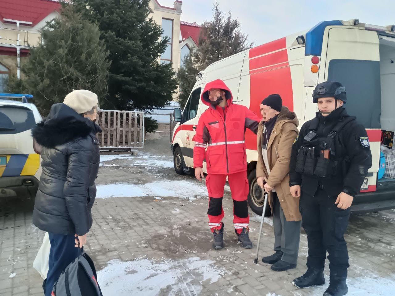 На фото зображено групу людей поруч із медичним автомобілем у зимовий час. Чоловік у червоному спецодязі рятувальника або медика спілкується з літньою людиною, яка спирається на тростину. Поруч стоїть представник поліції у захисному спорядженні, а ліворуч — жінка з рюкзаком. На фоні видно засніжене подвір'я та будівлі.