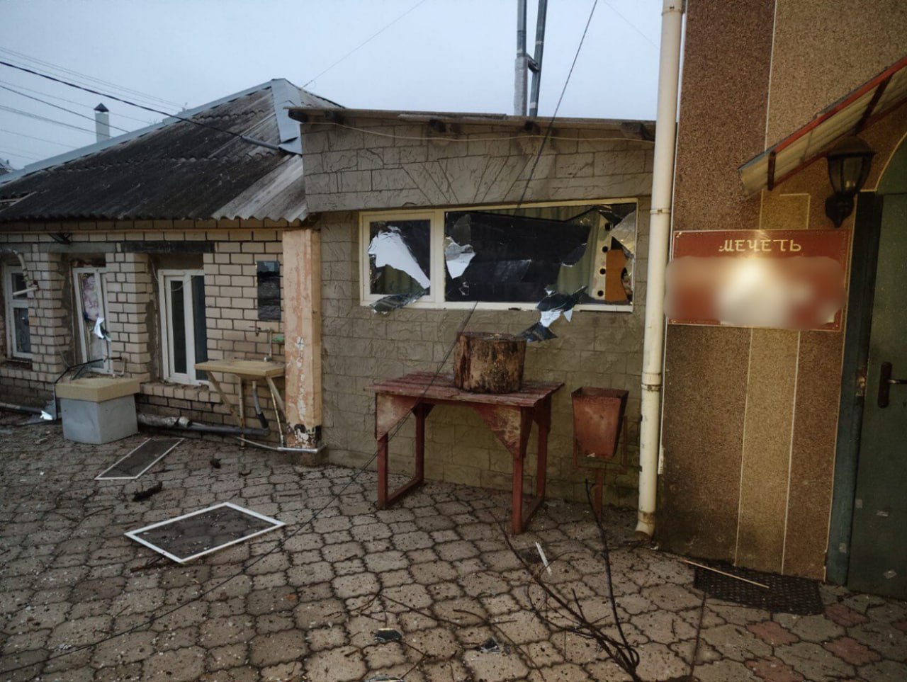 Фотографія наслідків обстрілу будівлі: вибиті шибки у вікнах, пошкоджені стіни та розкидане сміття на подвір'ї. На стіні біля входу розміщена табличка з написом «Мечеть».