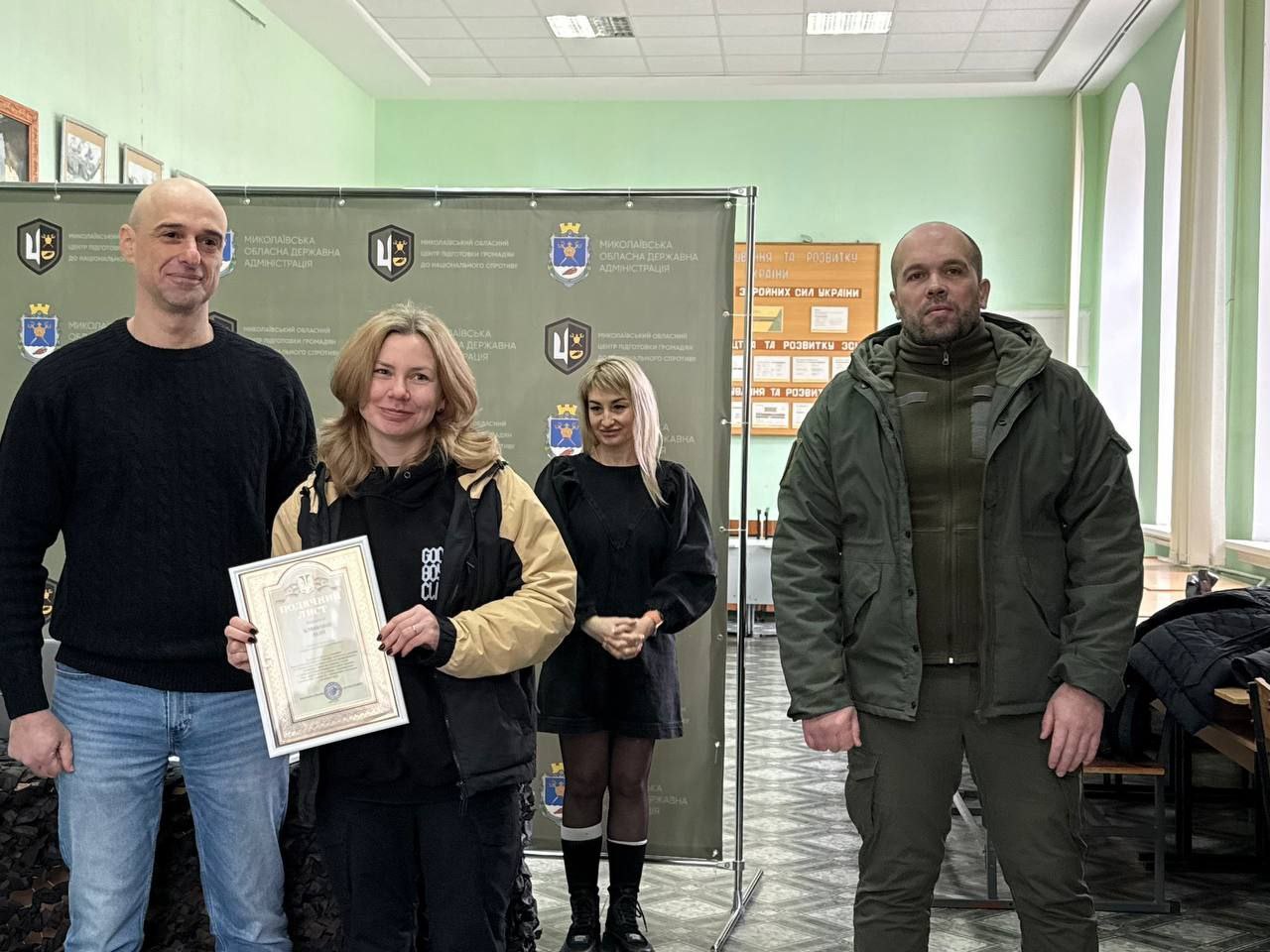 Жінка стоїть поруч із двома організаторами заходу, тримаючи в руках подяку в рамці. На задньому плані розташований банер з офіційною символікою, а праворуч на стіні — інформаційний стенд. Учасники одягнені у повсякденний та верхній одяг, атмосфера події офіційна та урочиста.