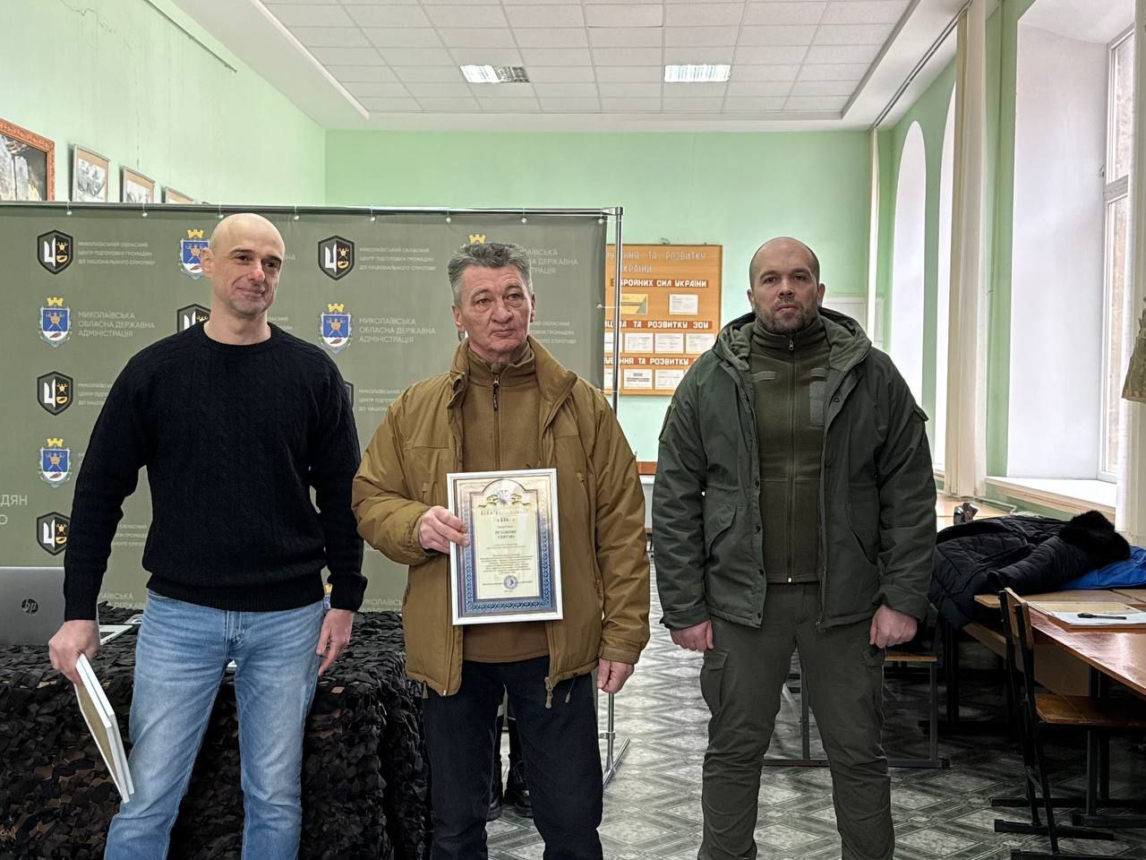 Чоловік у світло-коричневій куртці стоїть між двома організаторами заходу, тримаючи в руках почесну грамоту у рамці. Подія відбувається в офіційному приміщенні на фоні банера з державною символікою та патріотичних стендів. Учасники мають серйозний та урочистий вигляд.