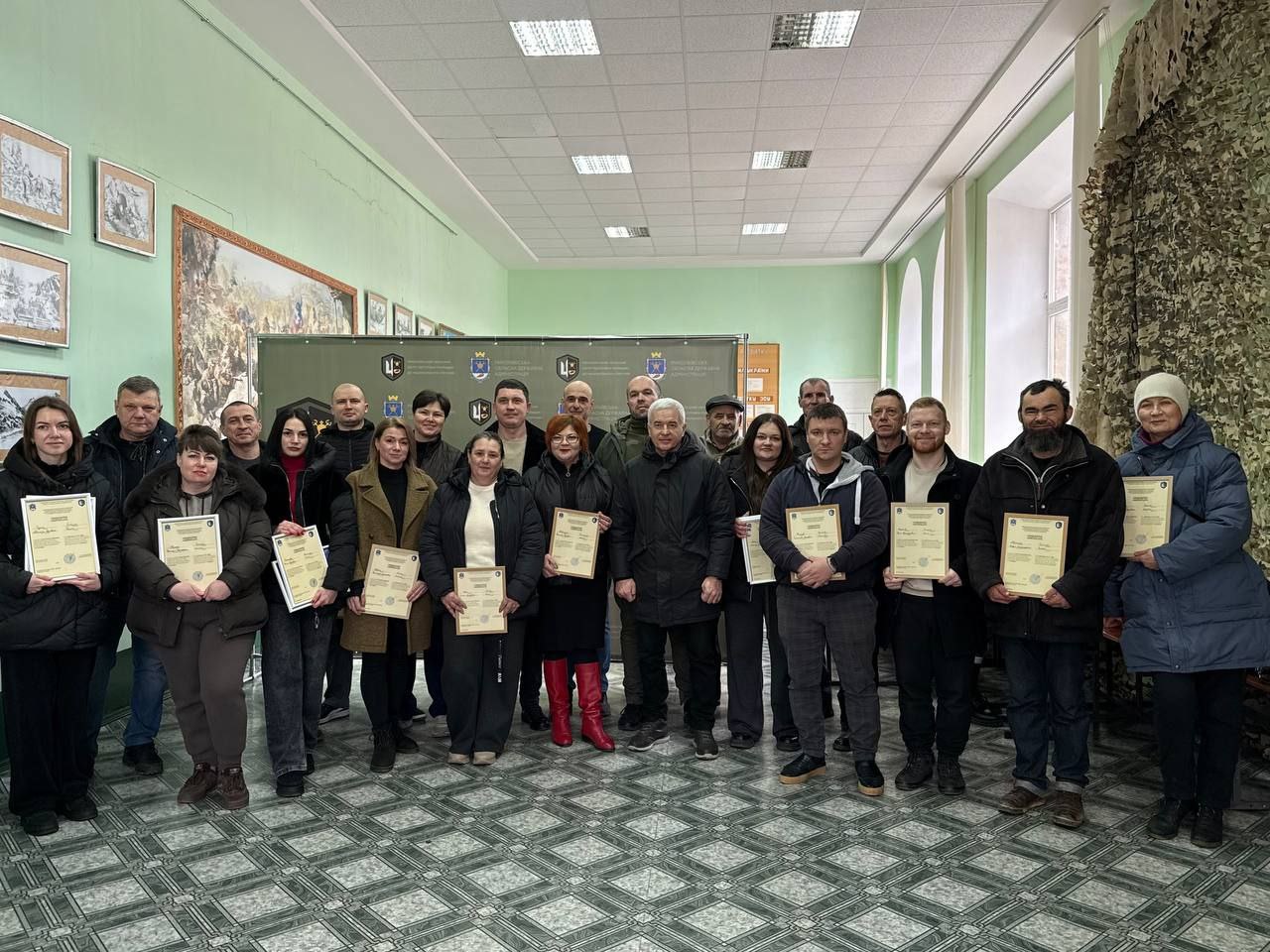 Група чоловіків та жінок стоїть у світлому коридорі офіційної установи, тримаючи в руках отримані сертифікати або подяки. На задньому плані розташований інформаційний стенд із державною символікою, а на стінах праворуч — патріотичні малюнки та маскувальна сітка. Учасники одягнені в теплий повсякденний одяг, атмосфера події виглядає урочистою.