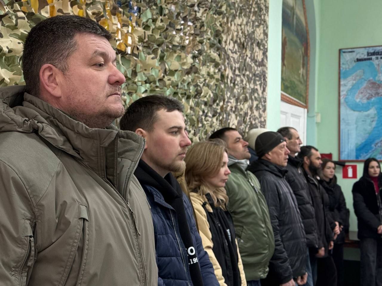 Група чоловіків та жінок у верхньому одязі стоїть у шерензі вздовж стіни в офіційному приміщенні. На задньому плані видно маскувальну сітку, патріотичні картини та велику карту на стіні. Люди стоять зосереджено, дивлячись в одному напрямку під час урочистої чи офіційної події.