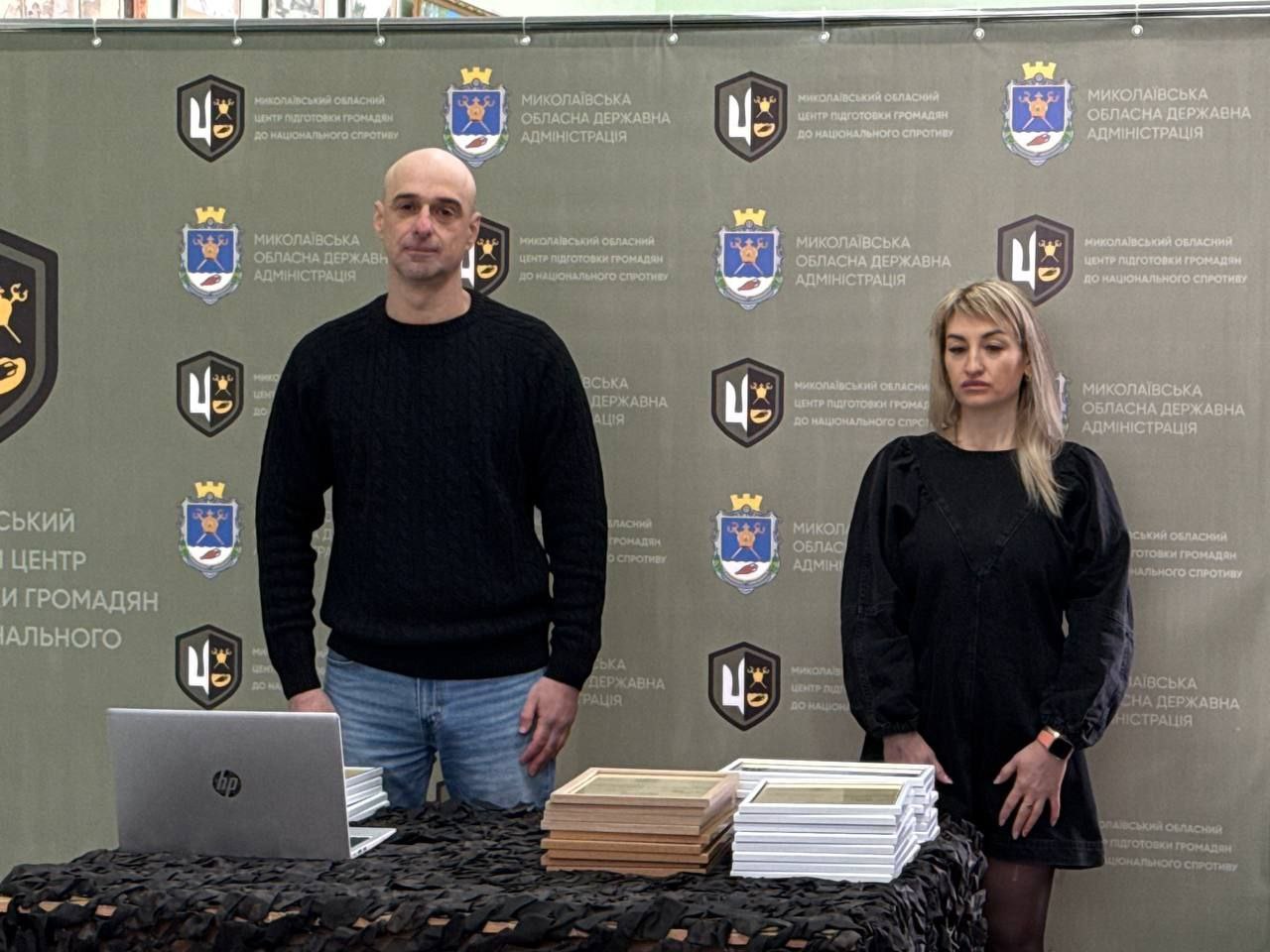 Чоловік та жінка стоять перед інформаційним банером із логотипами Миколаївської обласної державної адміністрації та Центру підготовки громадян до національного спротиву. Перед ними на столі, накритому маскувальною сіткою, лежать стоси готових сертифікатів або дипломів у рамках. Обоє одягнені в лаконічний темний одяг, зберігаючи офіційний та стриманий вигляд.