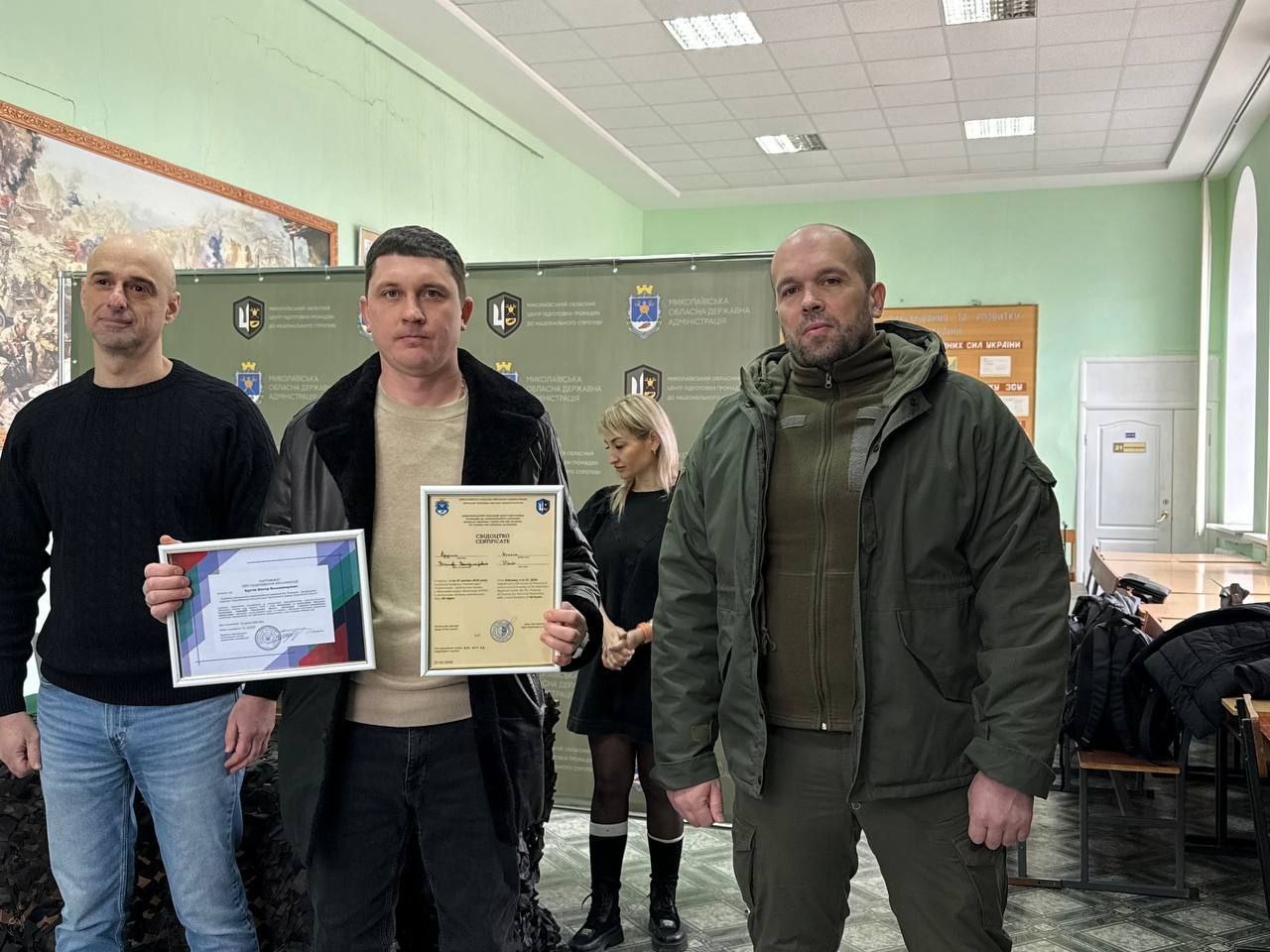 Троє чоловіків стоять разом у приміщенні на фоні офіційного банера з державною символікою. Один із них тримає в руках два сертифікати у рамках. Чоловіки одягнені у повсякденний та верхній одяг, на задньому плані видно частину навчального кабінету з патріотичними стендами.