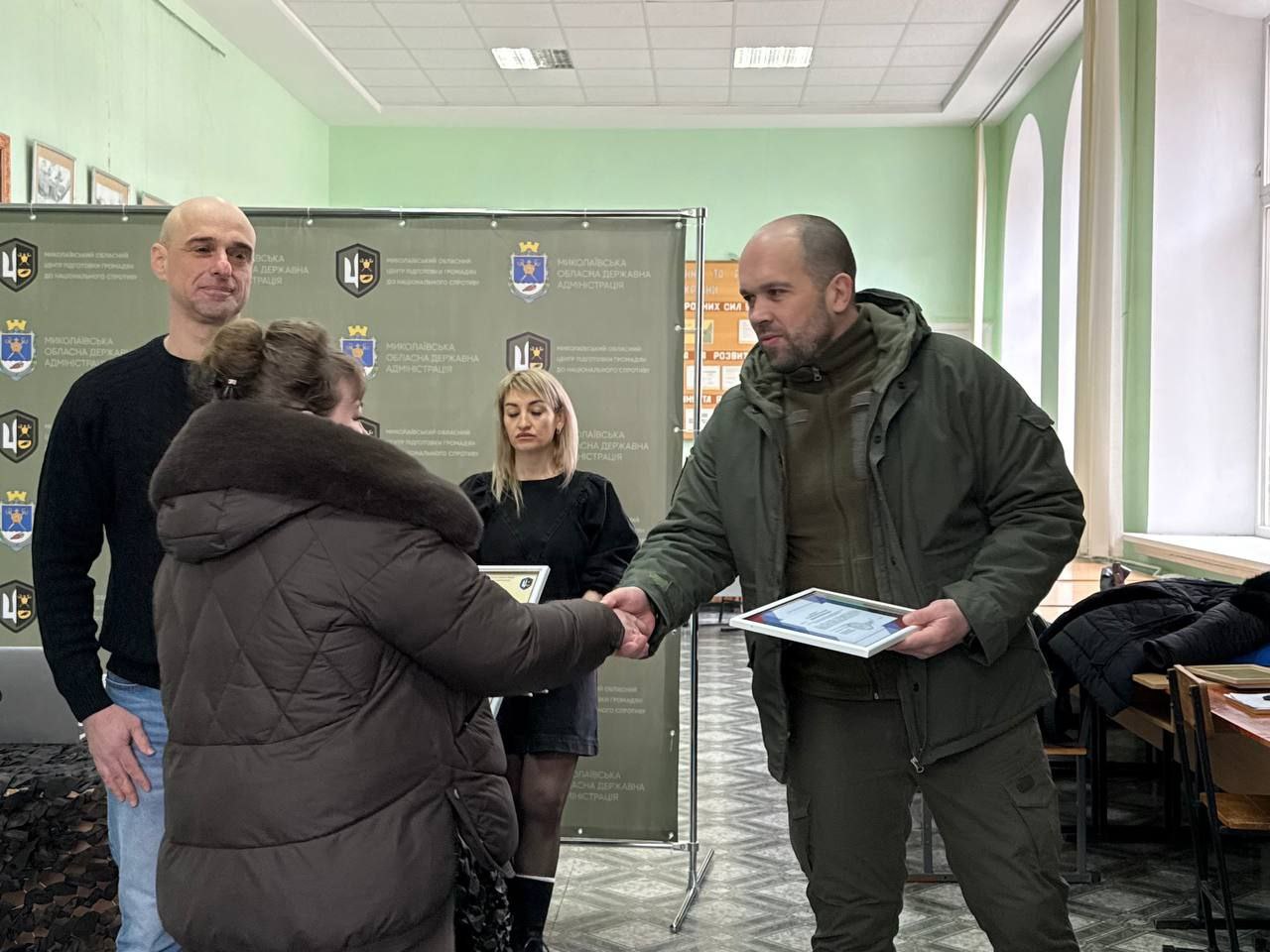 Чоловік у камуфляжній куртці тисне руку жінці, вручаючи їй сертифікат у рамці під час урочистої церемонії. Поруч стоять ще двоє організаторів заходу на фоні банера з офіційною символікою. Подія відбувається у просторому приміщенні з високими вікнами та патріотичними стендами на стінах.