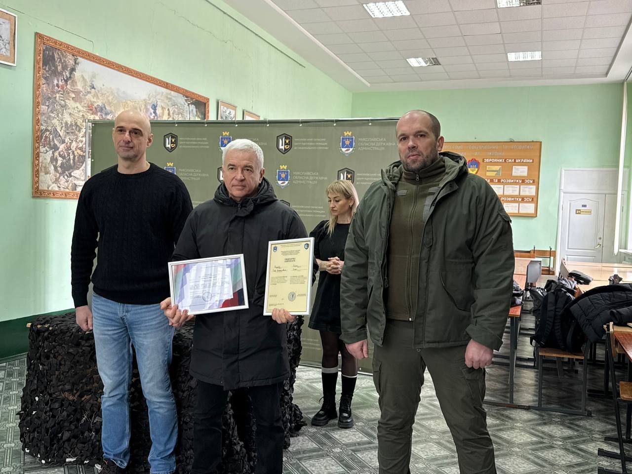 Чоловік у чорній куртці стоїть між двома іншими організаторами заходу, тримаючи в руках два офіційні сертифікати у рамках. Подія відбувається в аудиторії з патріотичними стендами на задньому плані.