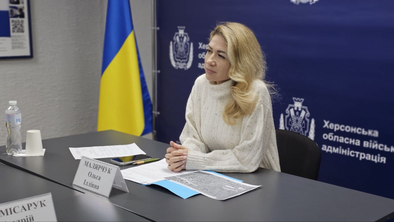 Жінка зі світлим волоссям у білому светрі сидить за столом під час офіційного заходу. Вона уважно слухає, склавши руки перед собою. На столі перед нею лежать папери, карта та табличка з ім'ям. Тло складає синій банер Херсонської обласної військової адміністрації та український прапор.