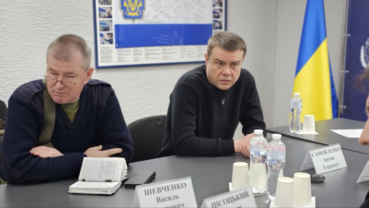 Двоє чоловіків сидять за столом під час наради. Один з них робить нотатки у блокноті, інший уважно дивиться вперед. На столі розміщені інформаційні таблички та пляшки з водою. На стіні позаду розміщено плакат із державною символікою, а поруч стоїть прапор України.