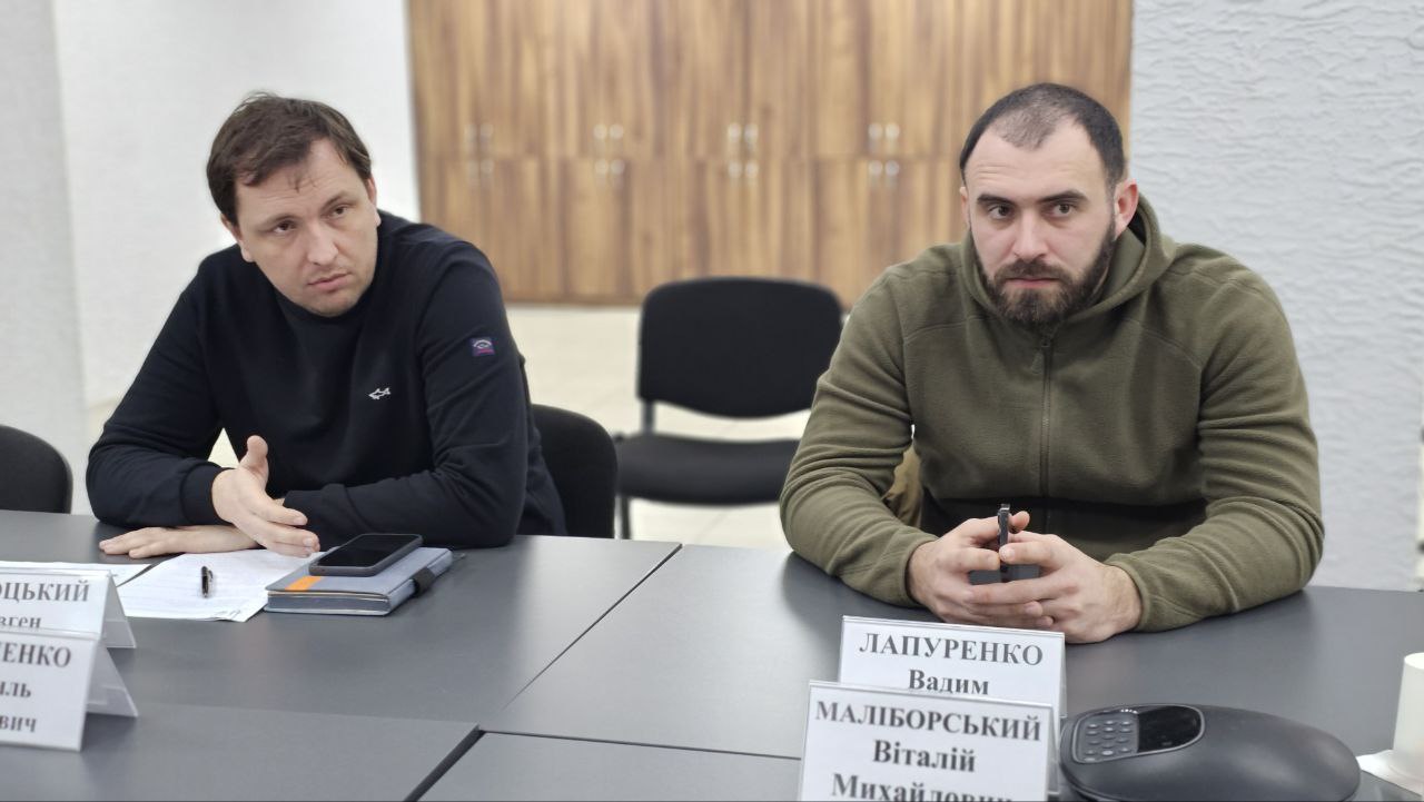 Двоє чоловіків у повсякденному одязі сидять за робочим столом під час засідання. Вони зосереджені на розмові, перед ними лежать документи та мобільні пристрої.