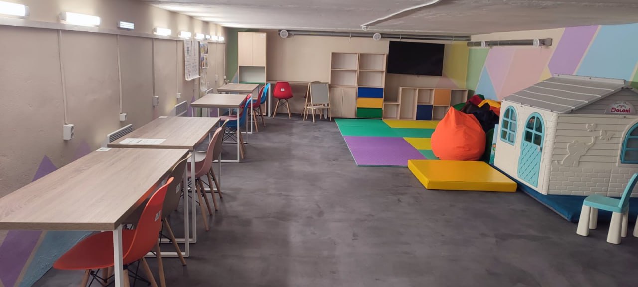 Облаштоване підвальне приміщення або укриття, розділене на зони для навчання та відпочинку. З одного боку стоять письмові столи з кольоровими стільцями, з іншого — дитячий ігровий будиночок, м'які крісла-мішки та кольорові мати на підлозі. Приміщення має світлі стіни з геометричними малюнками та гарне штучне освітлення.