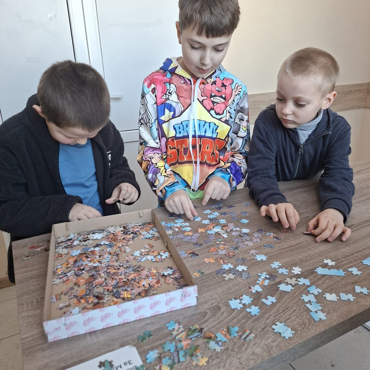 Троє хлопчиків зосереджено складають пазл за дерев'яним столом. Один із них вибирає деталі з коробки, а двоє інших розкладають їх на поверхні, намагаючись знайти правильні з'єднання. Діти одягнені у повсякденний теплий одяг, один із хлопчиків має яскраве худі з принтом. Процес виглядає спокійним та захопливим.