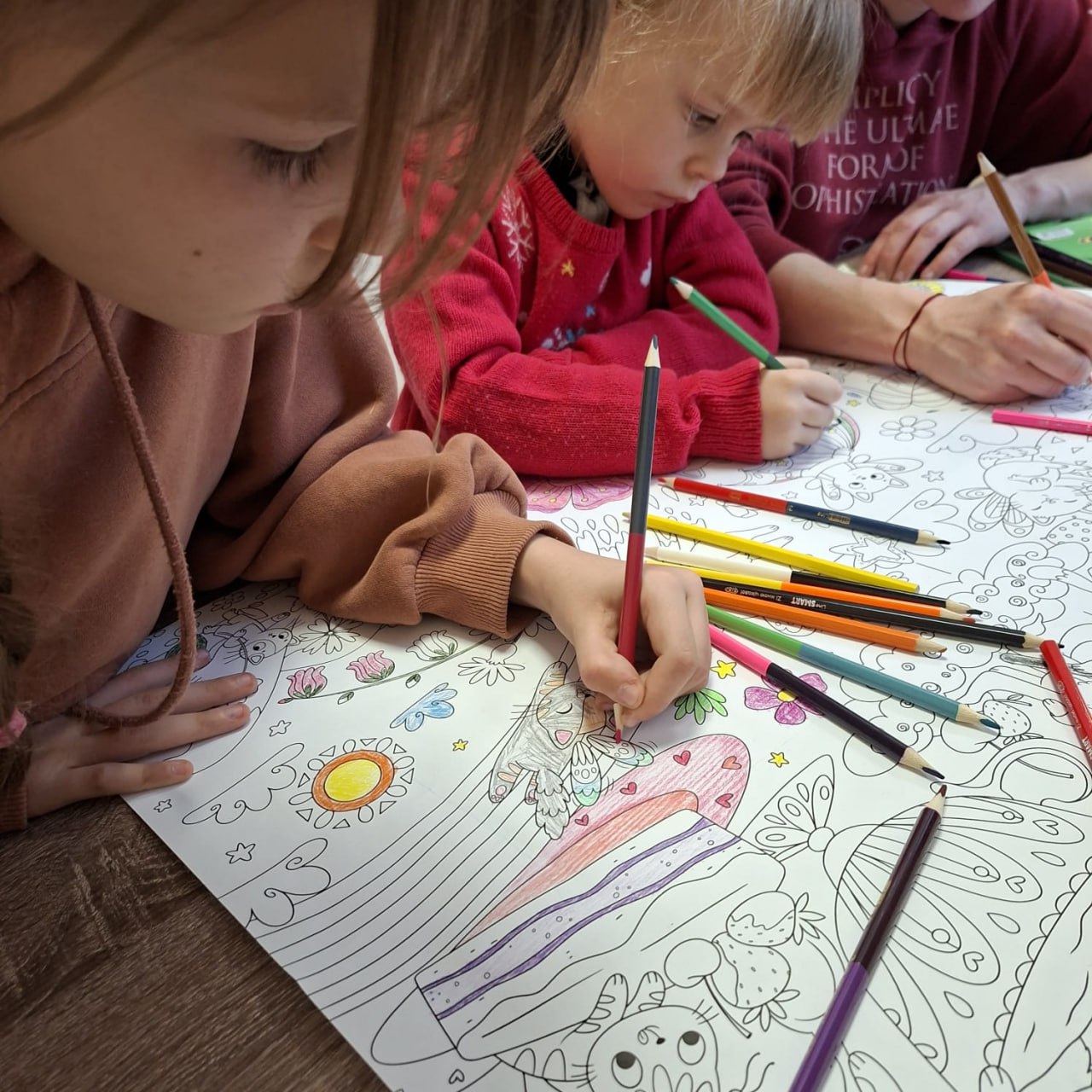Двоє маленьких дітей зосереджено розфарбовують велику паперову розмальовку кольоровими олівцями. На малюнку зображені квіти, комахи та сонце. Діти сидять за дерев'яним столом, поруч розкладено багато різнокольорових олівців, що свідчить про творчий процес у дружній атмосфері.