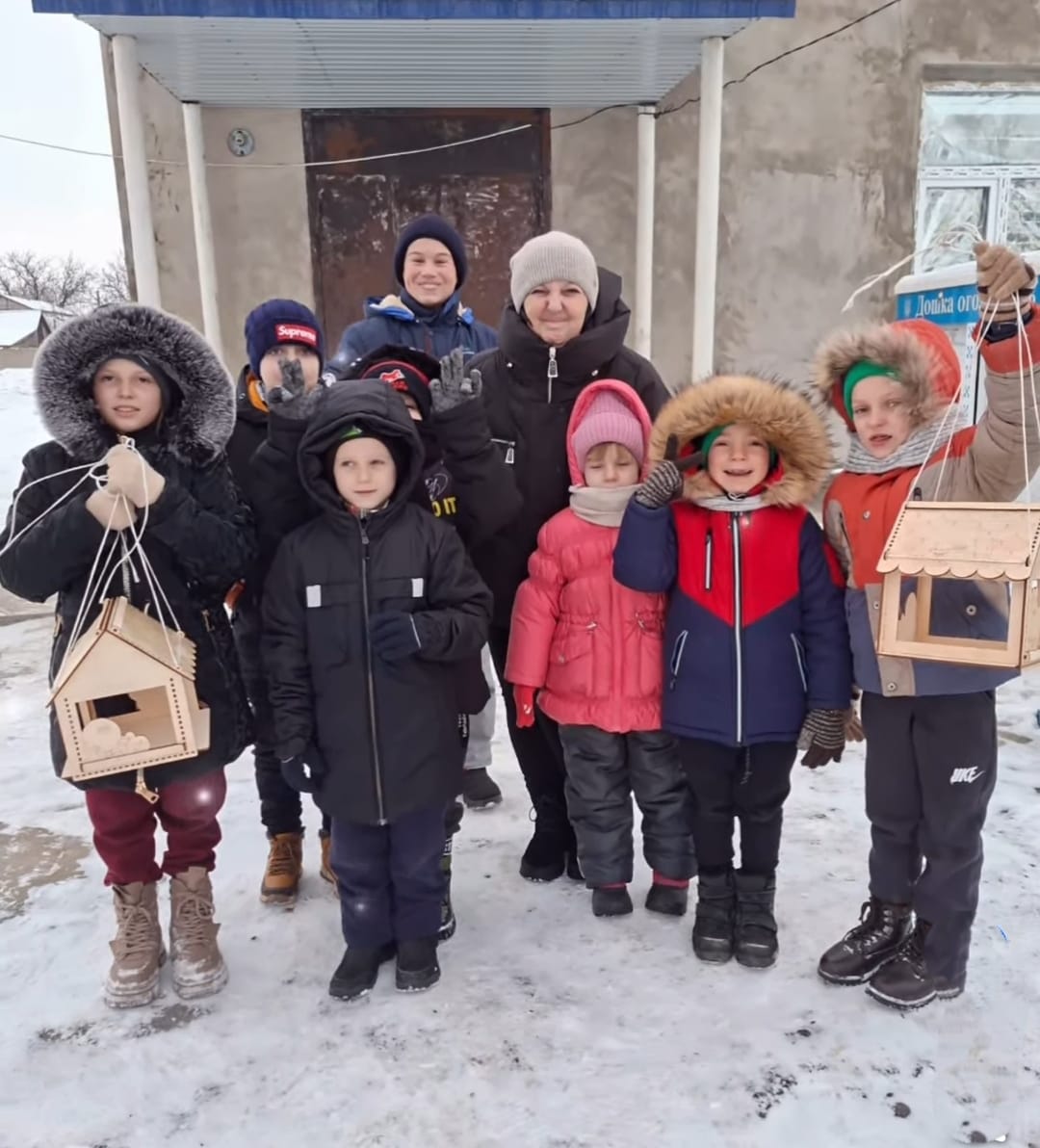 Група дітей різного віку разом із дорослою жінкою стоять на засніженому подвір'ї біля входу до будівлі. Діти одягнені в теплий зимовий одяг, двоє з них тримають у руках дерев'яні годівнички для птахів. Учасники виглядають задоволеними та позують для спільного фото.