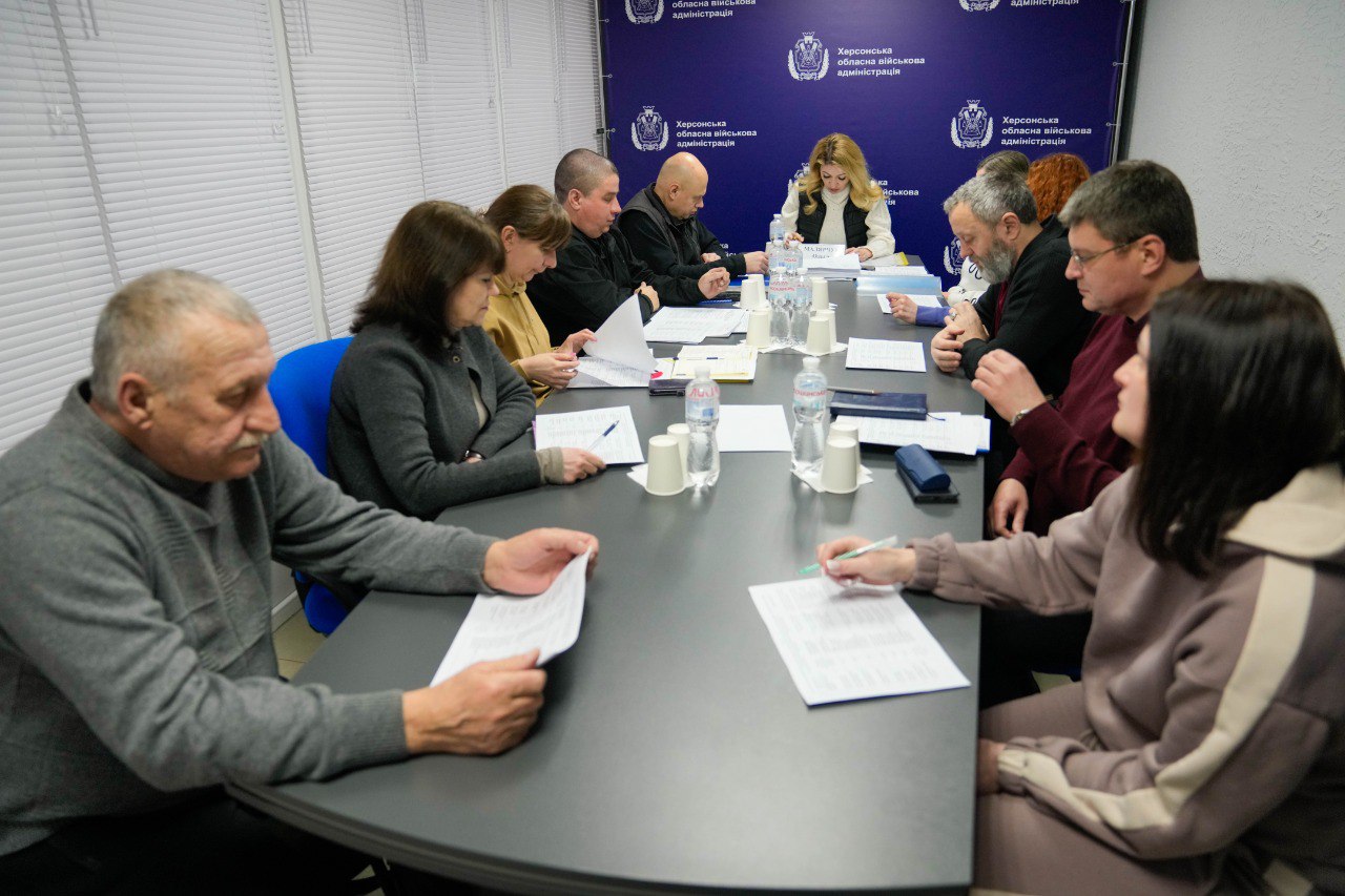 Робоча зустріч у залі засідань Херсонської ОВА. Учасники сидять за довгим столом, переглядаючи документи та ведучи обговорення. На задньому плані — синій офіційний банер установи та закриті жалюзі на вікнах.