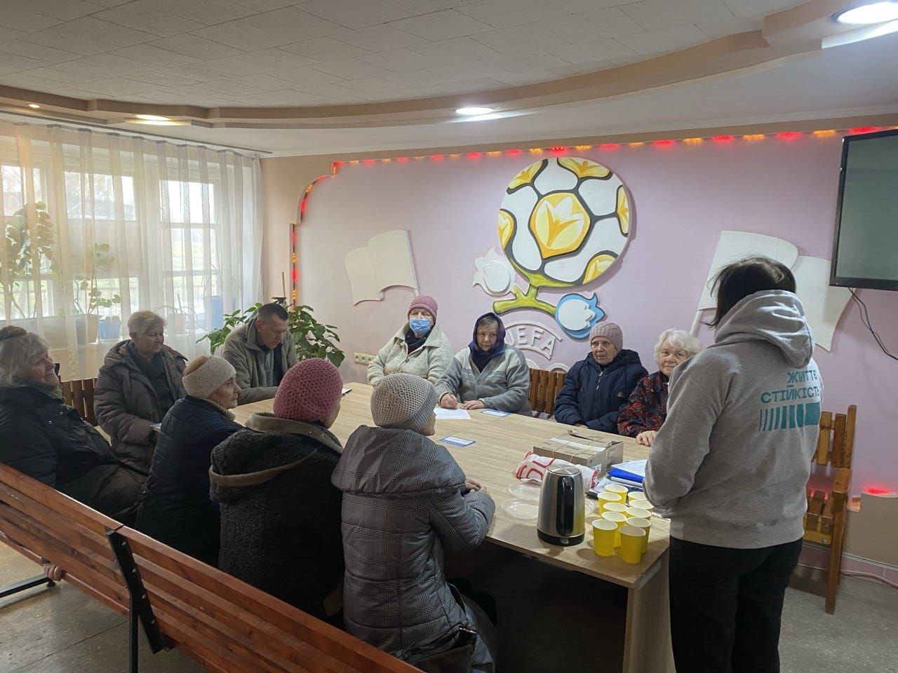 Група літніх людей сидить навколо довгого столу в приміщенні під час групової зустрічі. Перед ними стоїть ведуча в худі з написом «Життєстійкість». На столі розставлені паперові стаканчики та чайник, а на стіні позаду видно декоративну емблему та м'яке освітлення. Учасники одягнені в теплий верхній одяг та шапки, уважно слухаючи спікера.