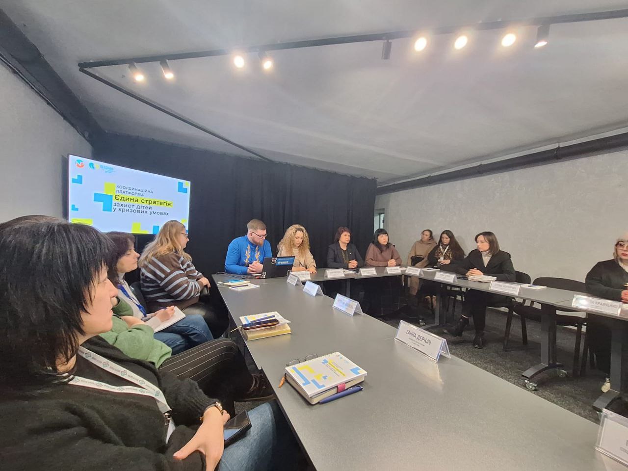 Група людей сидить за довгим столом під час офіційної зустрічі або круглого столу. На задньому плані видно екран із заголовком «Єдина стратегія: захист дітей у кризових умовах». Учасники заходу зосереджено слухають або готуються до обговорення, на столі перед ними лежать блокноти та ноутбук.