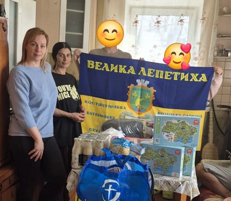 Група людей стоїть у приміщенні біля столу з гуманітарною допомогою (продуктами та іграшками). Вони тримають синьо-жовтий прапор із написом «Велика Лепетиха» та назвами інших населених пунктів.