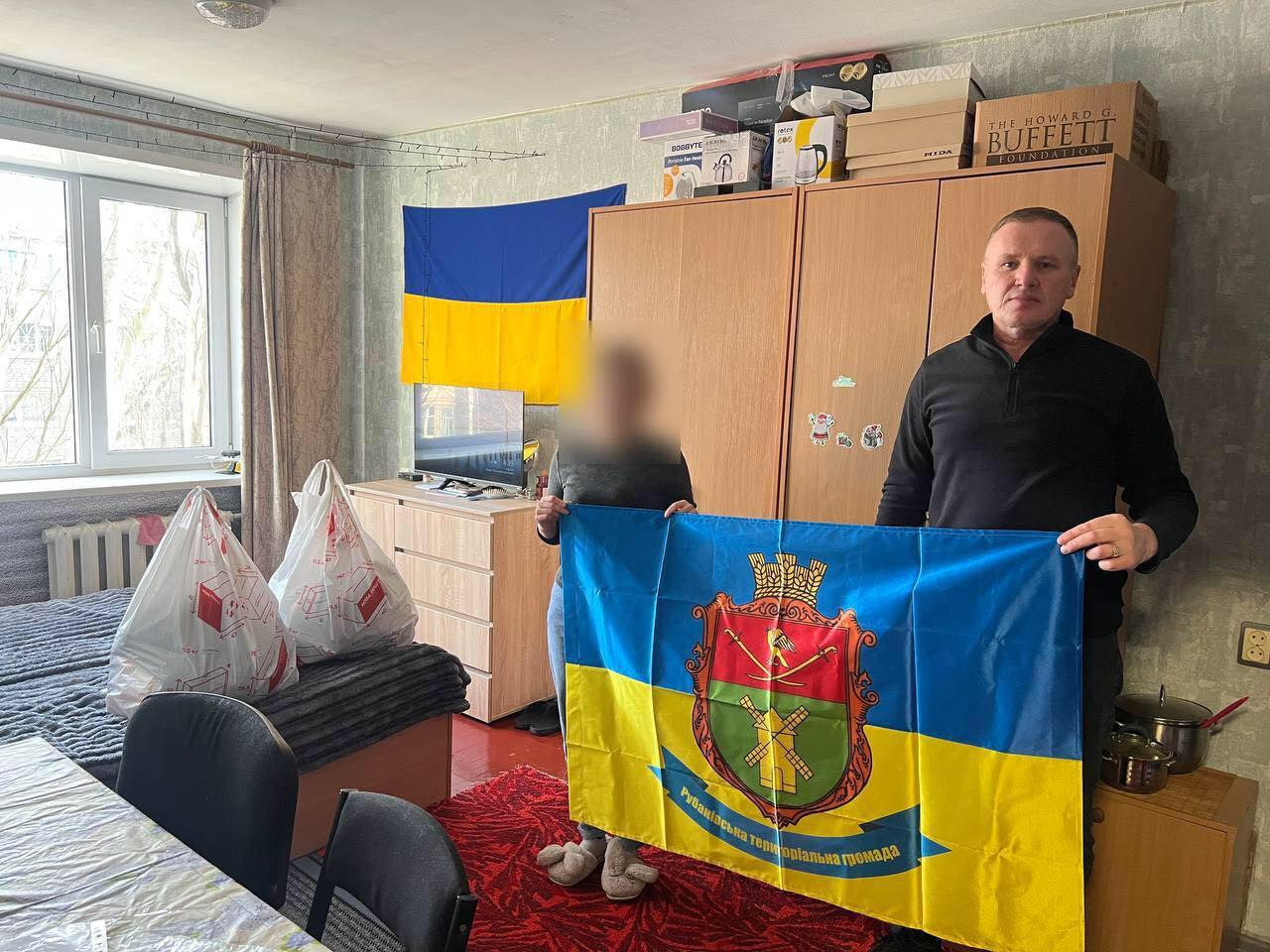 Двоє людей у житловій кімнаті тримають прапор Рубанівської територіальної громади. На задньому плані видно державний прапор України, побутові речі та пакунки з гуманітарною допомогою.