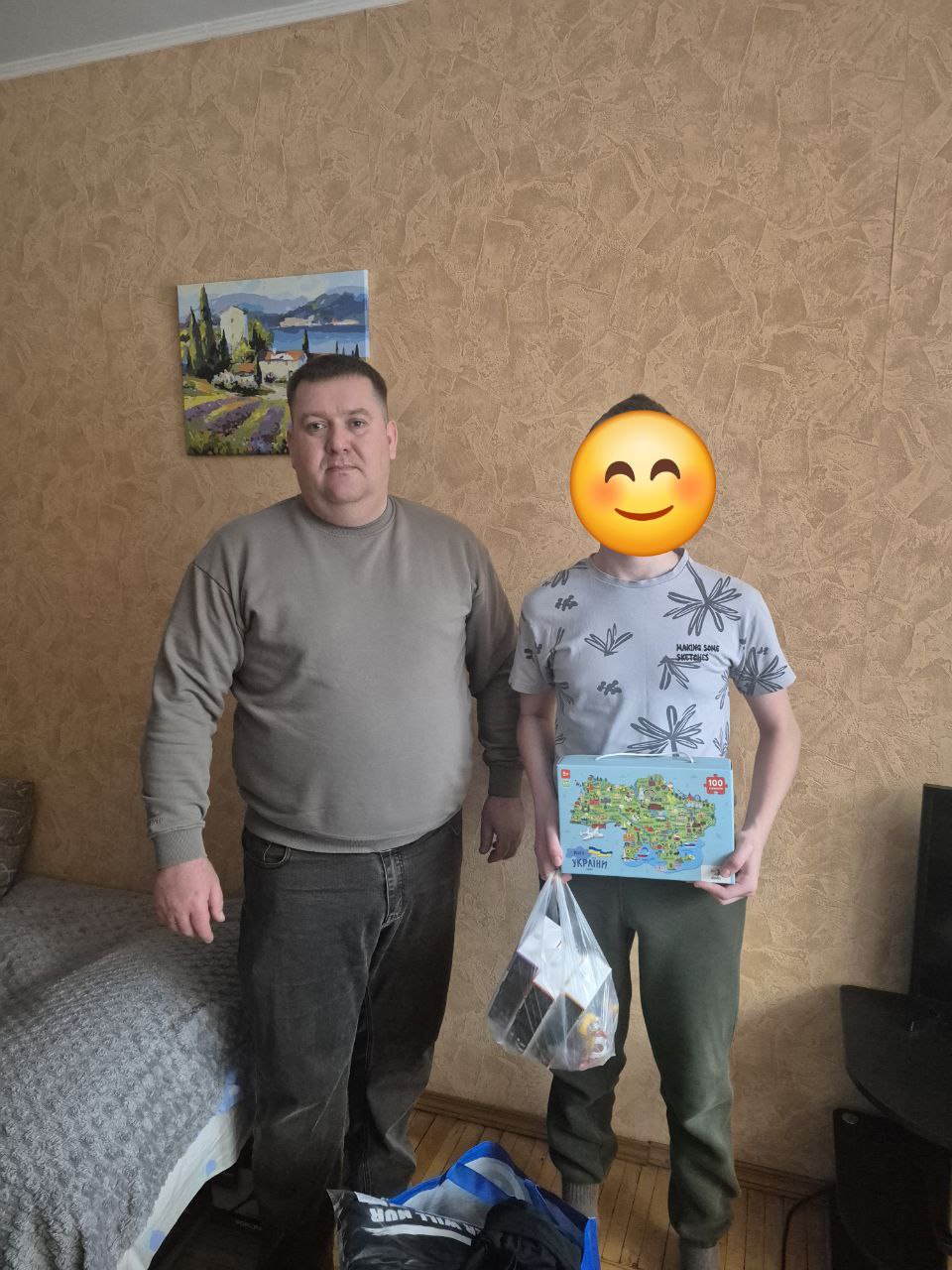Двоє людей стоять у кімнаті. Один із них тримає в руках коробку з пазлом у формі карти України та пакет із речами. Поруч на підлозі стоїть велика сумка з допомогою.