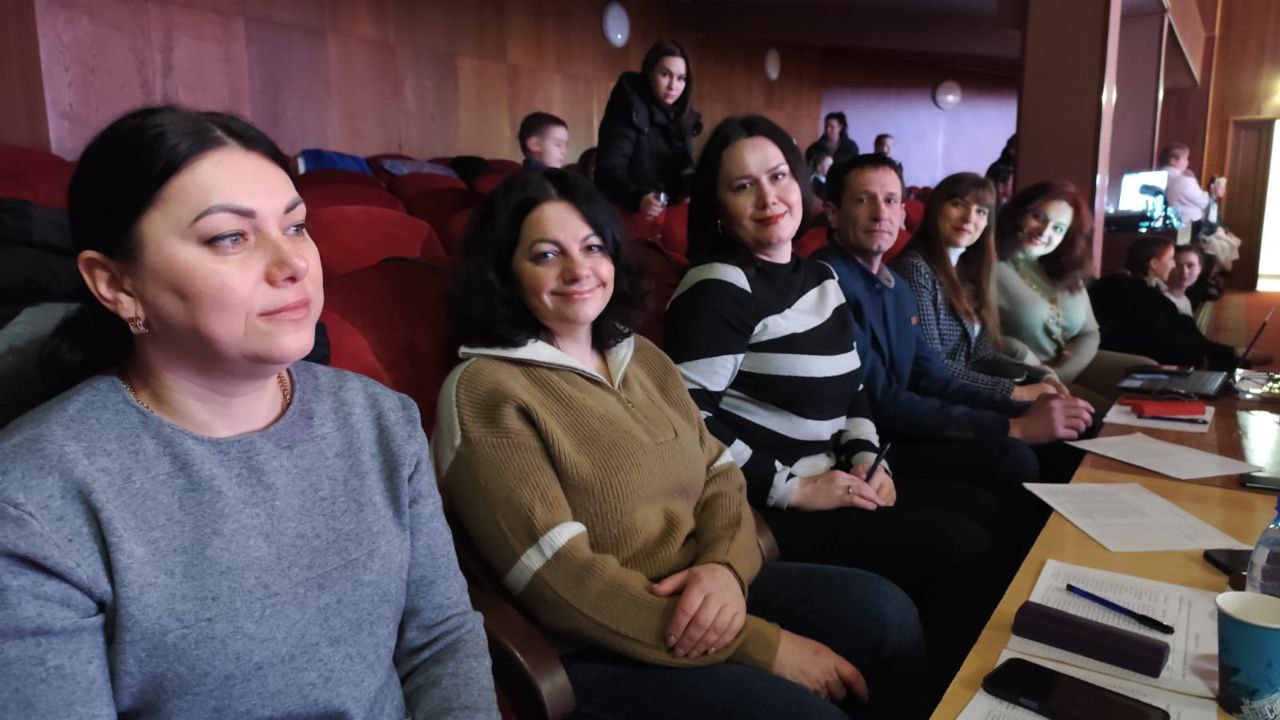 Група людей сидить у глядацькій залі під час заходу. На передньому плані видно членів журі або організаторів, які сидять за столом із документами, а на задньому плані — глядацькі крісла.